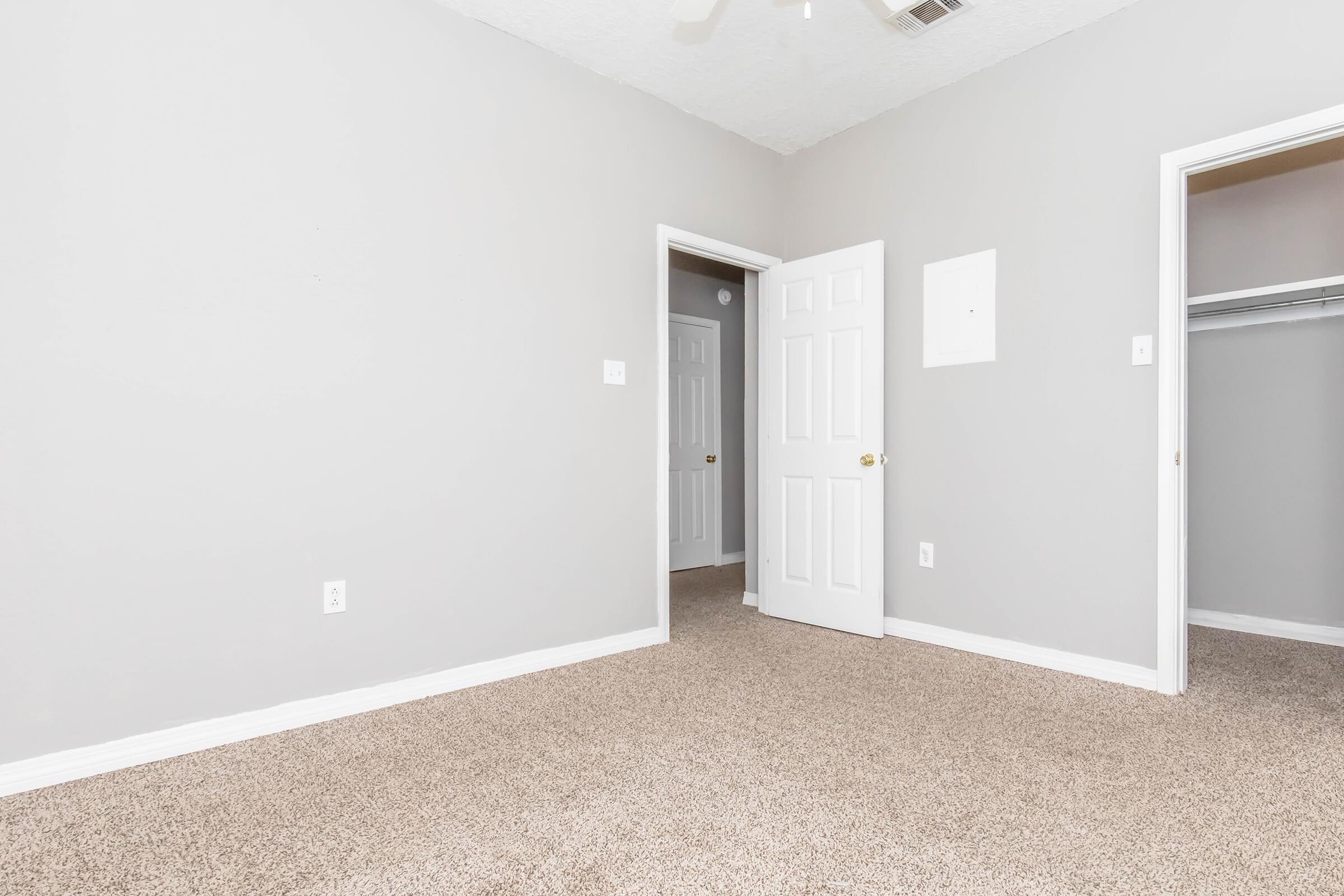 A simple, unfurnished room with light gray walls and beige carpet. There are two doors leading to other rooms, one partially open, and a single switch plate on the wall. The space is bright and well-lit, creating a clean and inviting atmosphere.