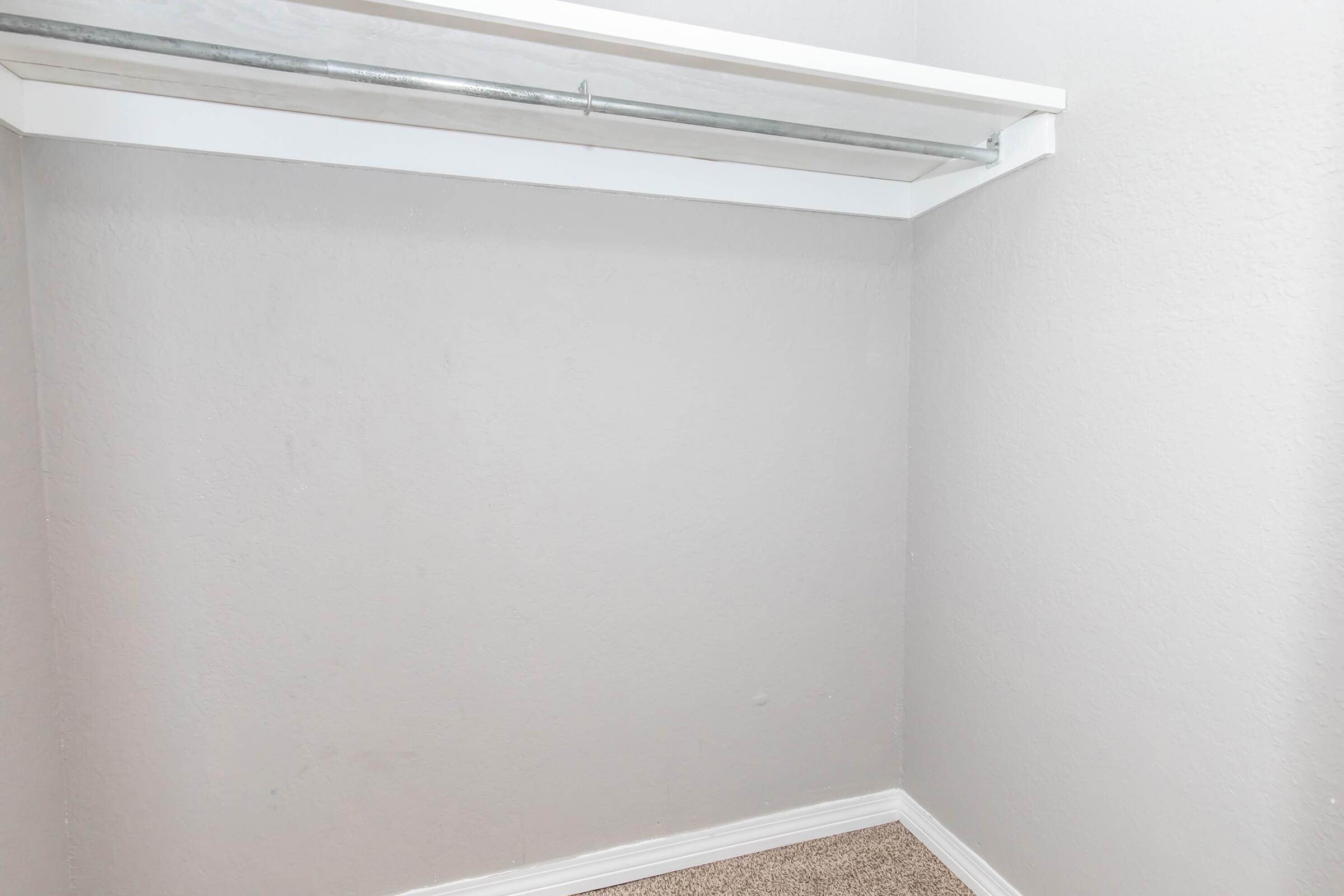Empty closet with light gray walls and a white shelf. The floor is carpeted, and there is a metal rod for hanging clothes mounted along the top. The space appears tidy and uncluttered.