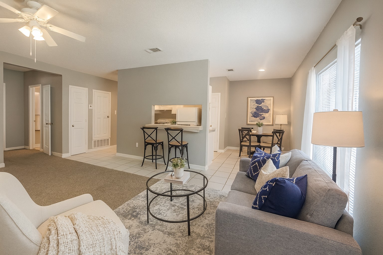 A cozy and modern living room featuring a light gray sofa with decorative pillows, a circular coffee table, and a stylish armchair. There is a small dining area with two chairs, a light-colored wall, and a ceiling fan overhead. Large windows let in natural light, enhancing the inviting atmosphere.