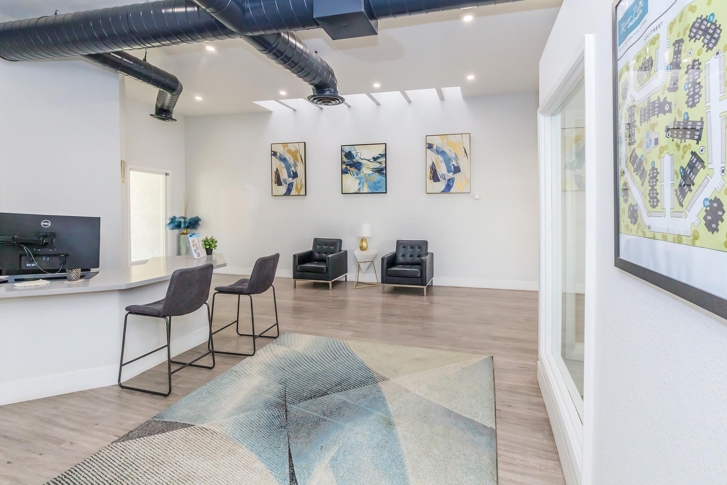 A modern reception area featuring a sleek desk with a computer, two black armchairs, and three abstract wall art pieces. The flooring is wooden, and a large area rug with a geometric design is visible. Natural light fills the space through a skylight, creating a welcoming atmosphere.