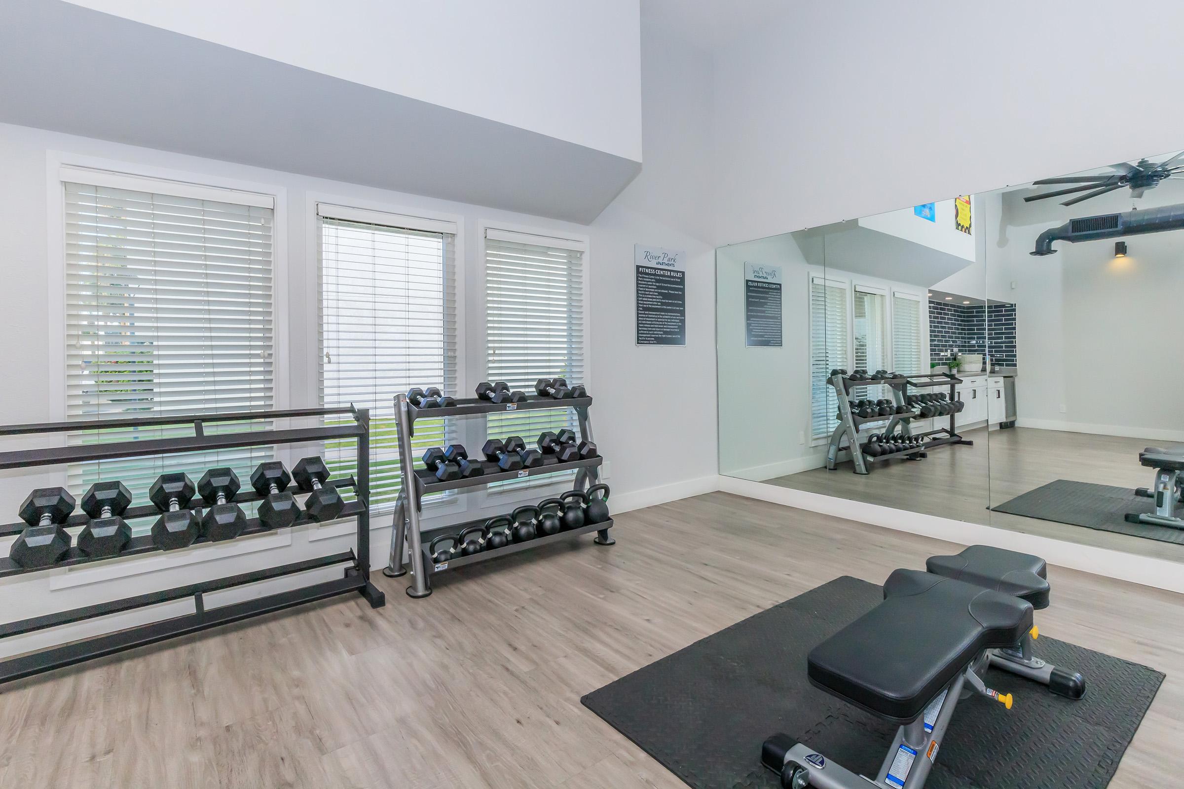 A well-lit home gym featuring a mirrored wall, a weight bench, and a rack of dumbbells. Natural light enters through large windows, and the floor is a light wood or laminate finish. The space is clean and organized, with exercise equipment neatly arranged, creating a motivating workout environment.
