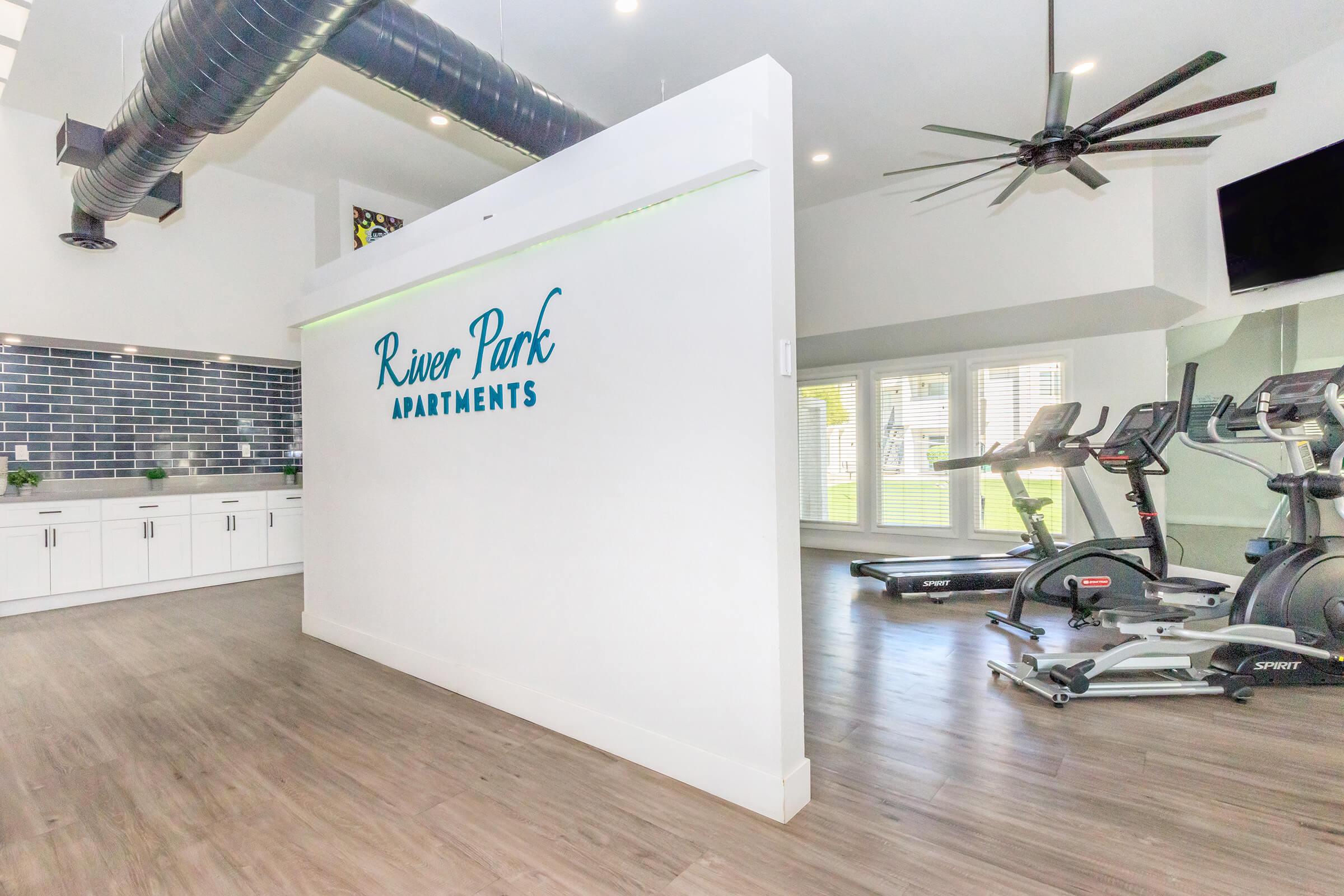 Interior view of River Park Apartments featuring a modern fitness area. A white wall with the text "River Park Apartments" is prominent, and there's a kitchenette area with cabinets. To the right, there are exercise machines, large windows providing natural light, and ceiling fans above.