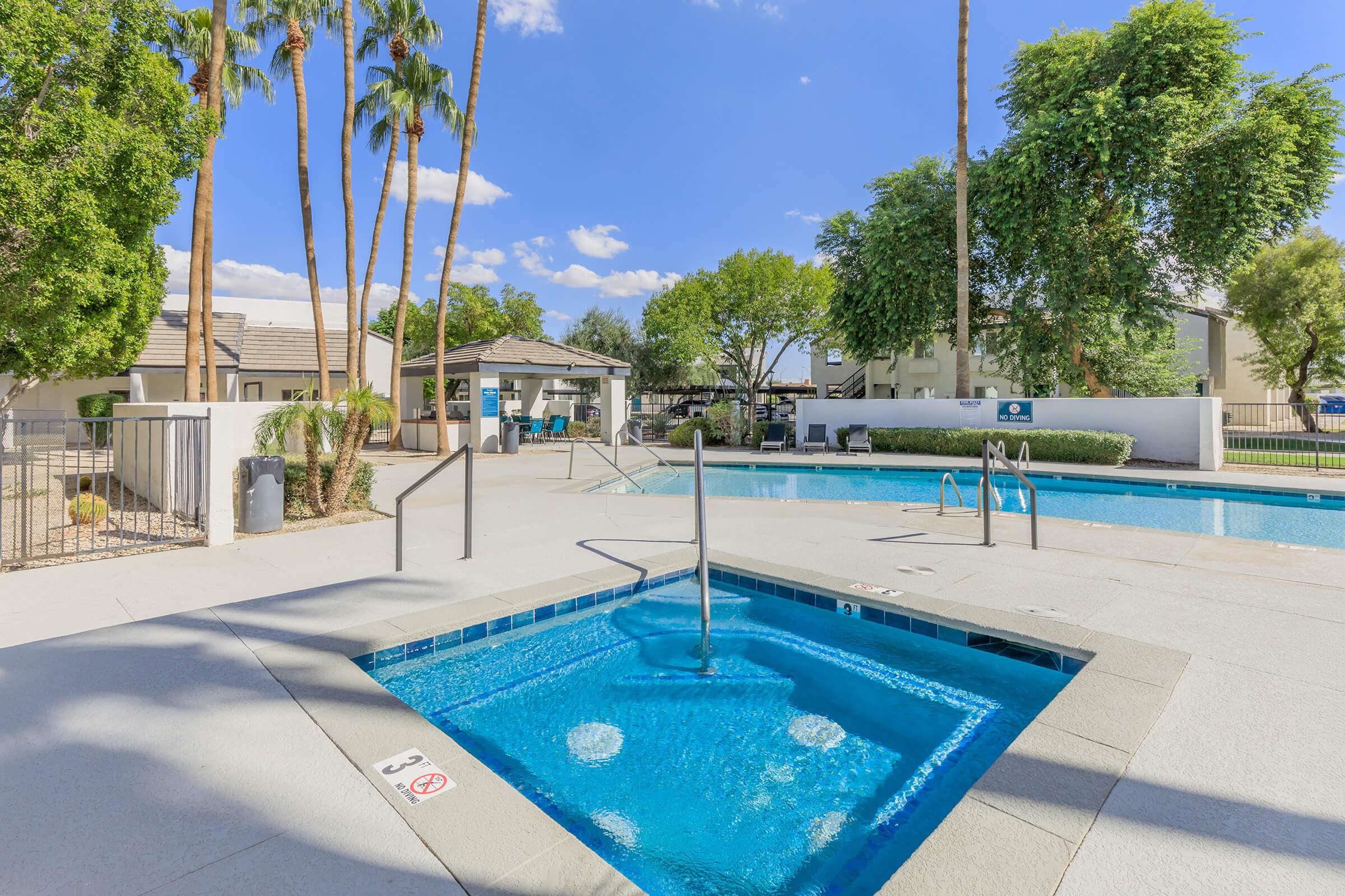 A sunny outdoor pool area with palm trees and lush greenery. The image features two swimming pools, one of which has a jacuzzi. There are lounge chairs and a shaded gazebo nearby, with blue skies and fluffy clouds in the background, creating a relaxing atmosphere.