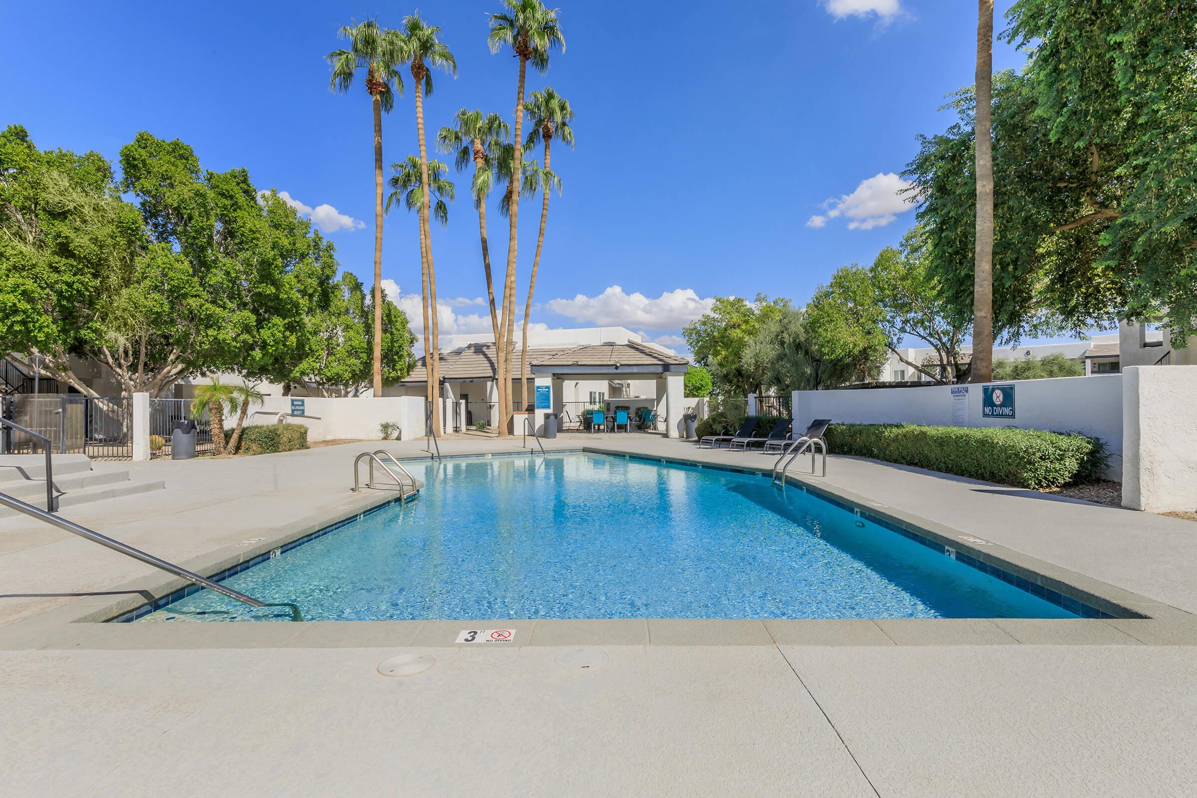 A clear swimming pool surrounded by palm trees, with a small seating area and clubhouse visible in the background. The sky is bright blue with a few fluffy clouds, creating a serene outdoor atmosphere. The pool area is well-maintained and inviting.