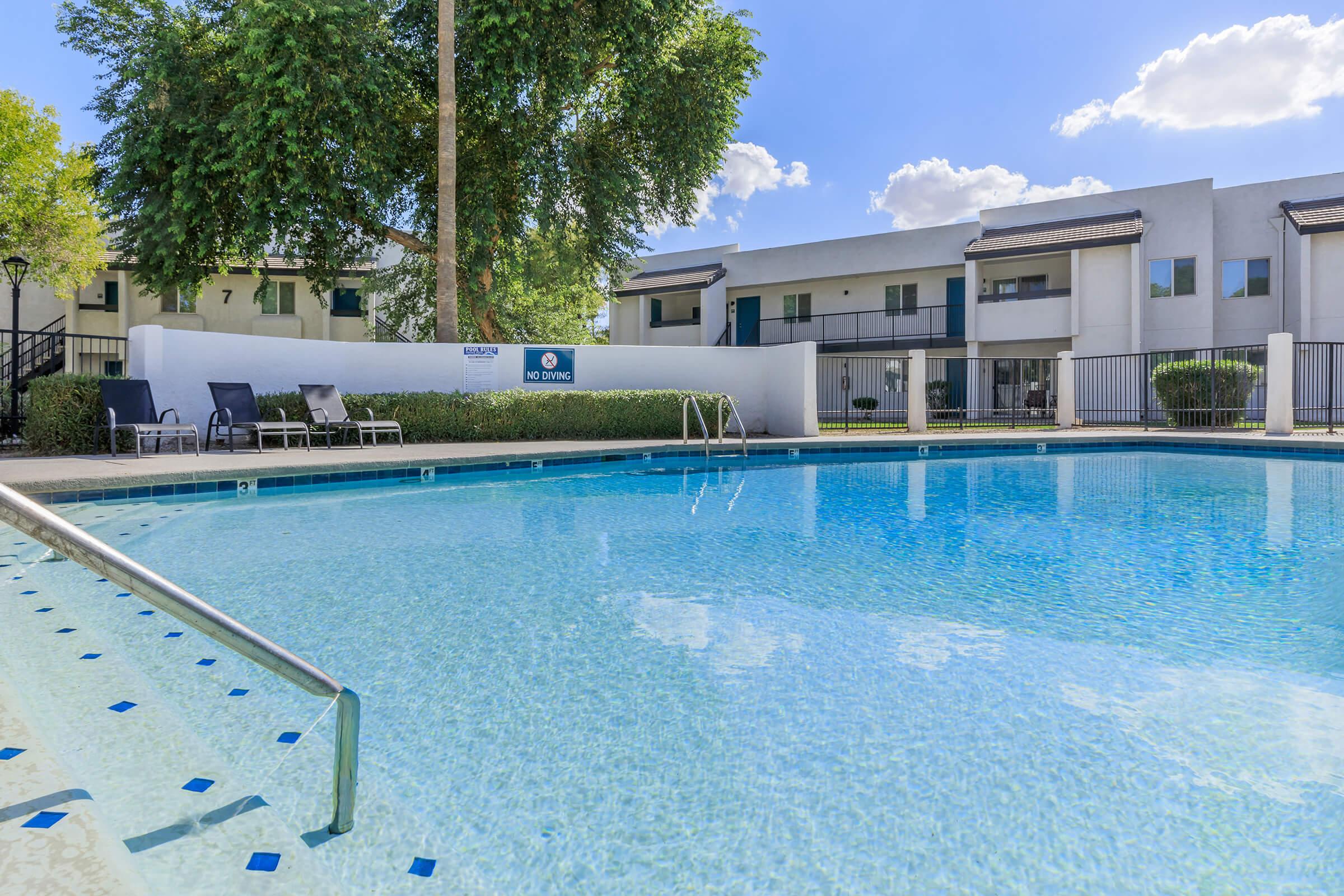 A clear blue swimming pool surrounded by lounge chairs and trees, with a white building in the background. The pool area is inviting, featuring a no diving sign, under a bright sky with fluffy clouds.
