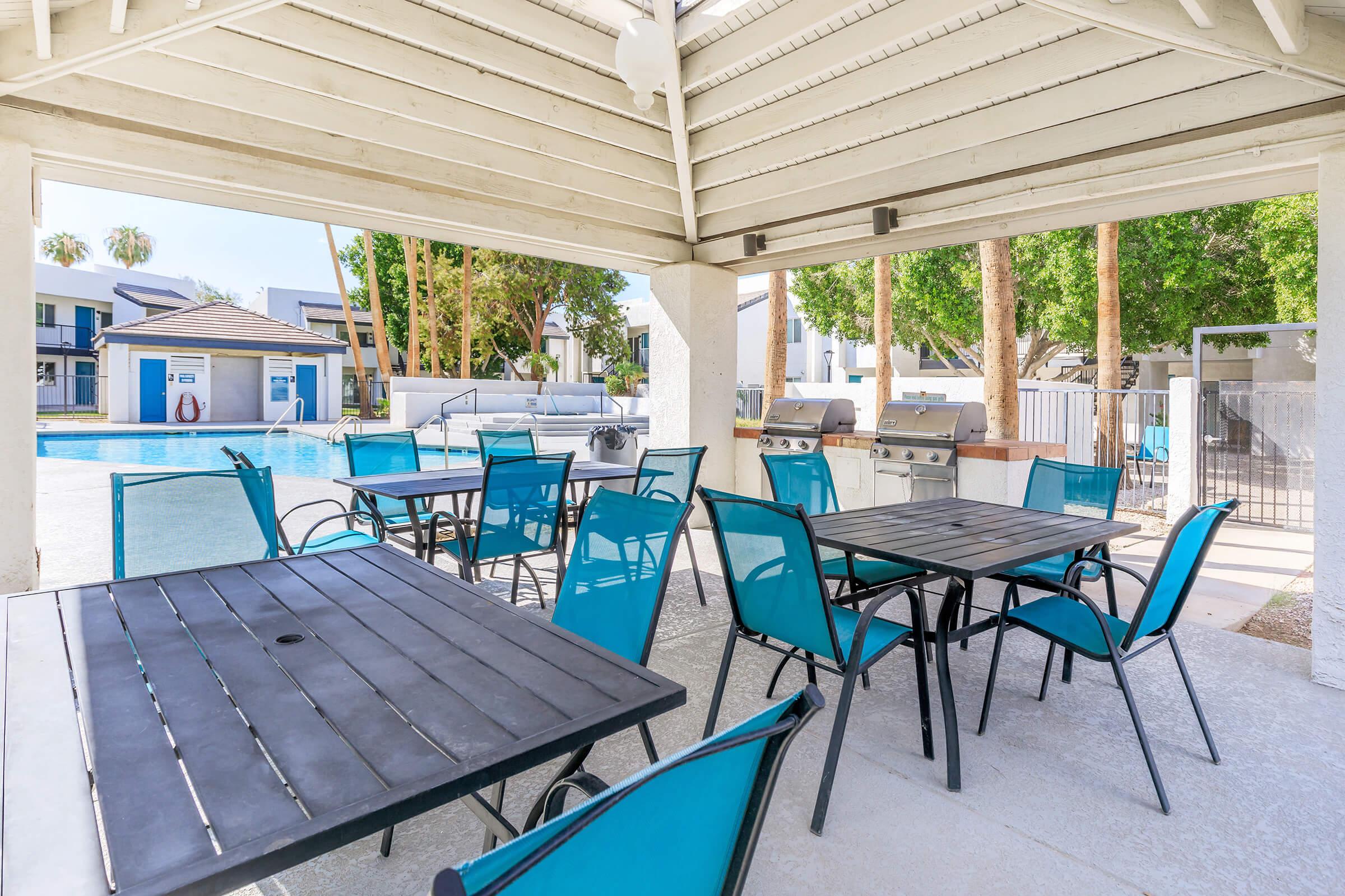 Outdoor seating area with turquoise chairs and black tables under a shaded pavilion. In the background, a swimming pool is visible, along with grill stations. Lush green trees surrounding the area create a relaxing atmosphere. Ideal for social gatherings or leisurely afternoons by the pool.