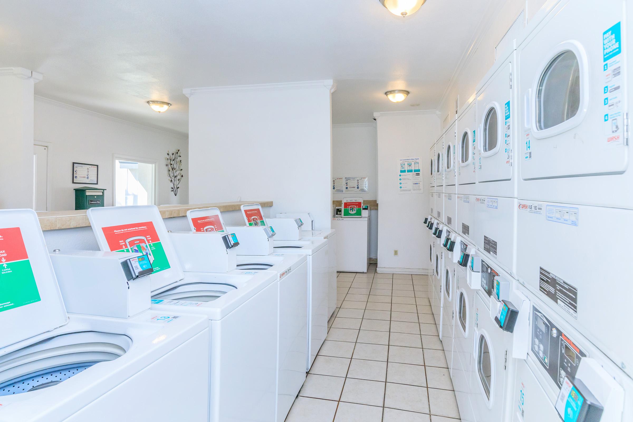 A brightly lit laundromat featuring a row of white washing machines and dryers. The machines have digital displays and are neatly arranged in a spacious, clean environment. The floor is tiled, and the walls are painted white, creating a fresh and inviting atmosphere.