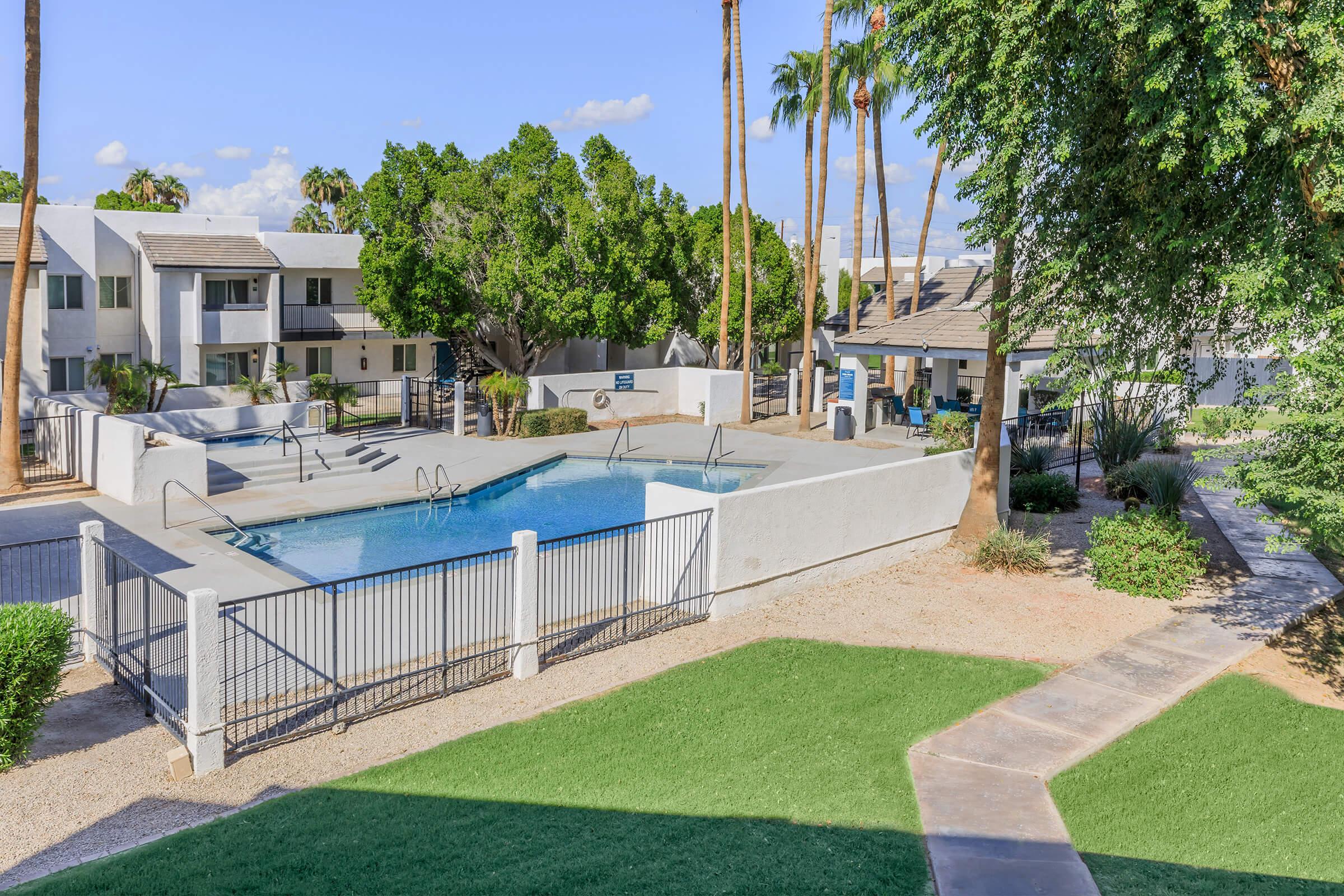 A serene view of an apartment complex featuring a blue swimming pool surrounded by palm trees and landscaped greenery. The pool area includes metal fencing and lounge chairs, with nearby pathways and buildings visible in the background under a clear blue sky.