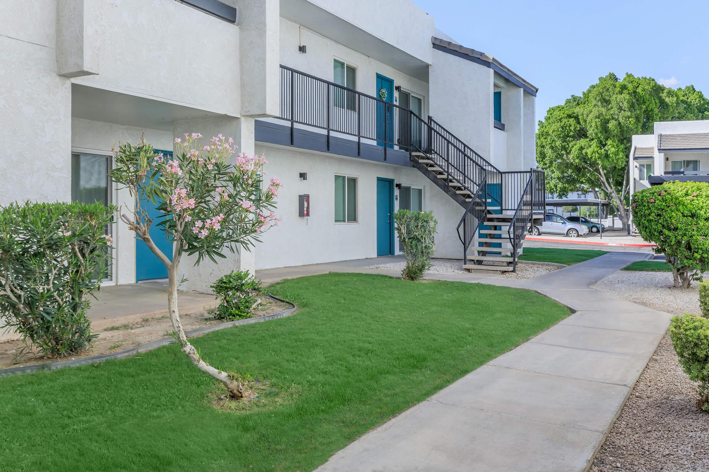 A view of a modern apartment complex featuring two-story buildings with blue doors, surrounded by well-maintained greenery. The pathway leads to a staircase, and there are sandy areas and shrubs in the landscape, creating a welcoming outdoor space.