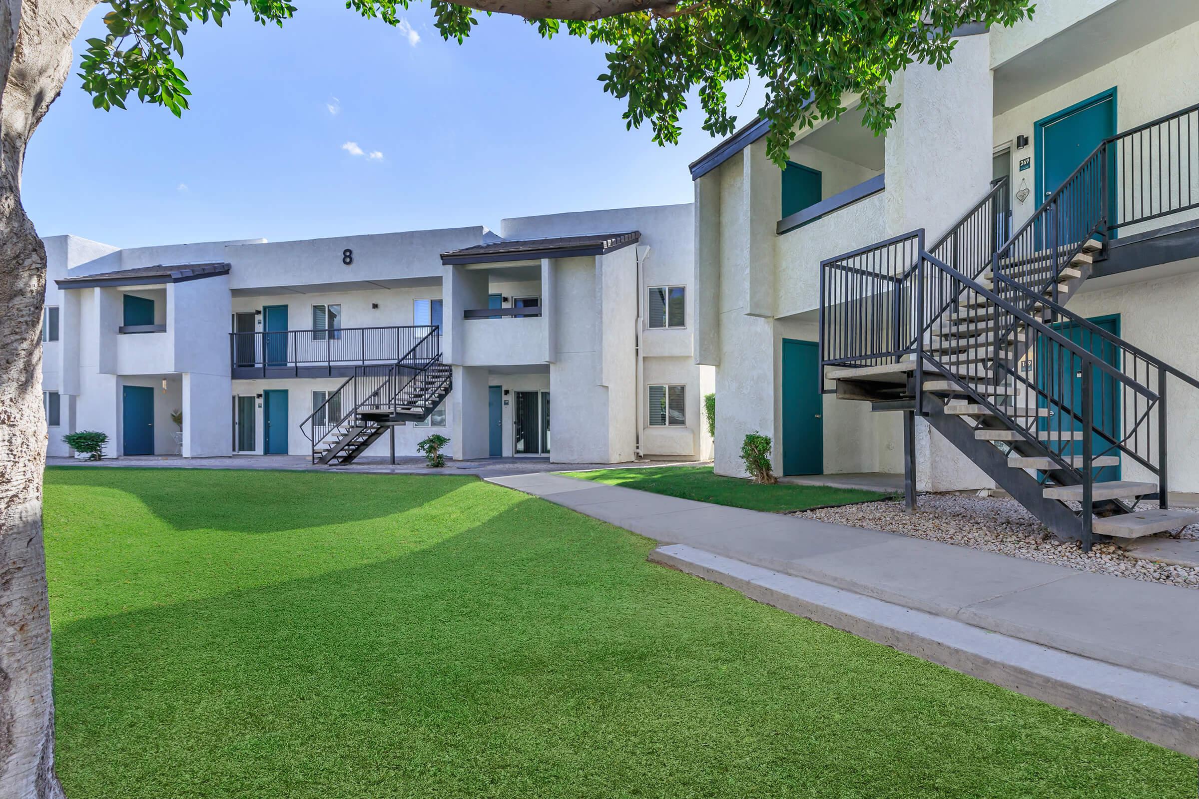 A modern apartment complex featuring three buildings with white exteriors and green doors. There are metal stairs leading to the upper floors. The landscape includes manicured green grass and a tree, with a walkway connecting the buildings. The sky is clear and blue.