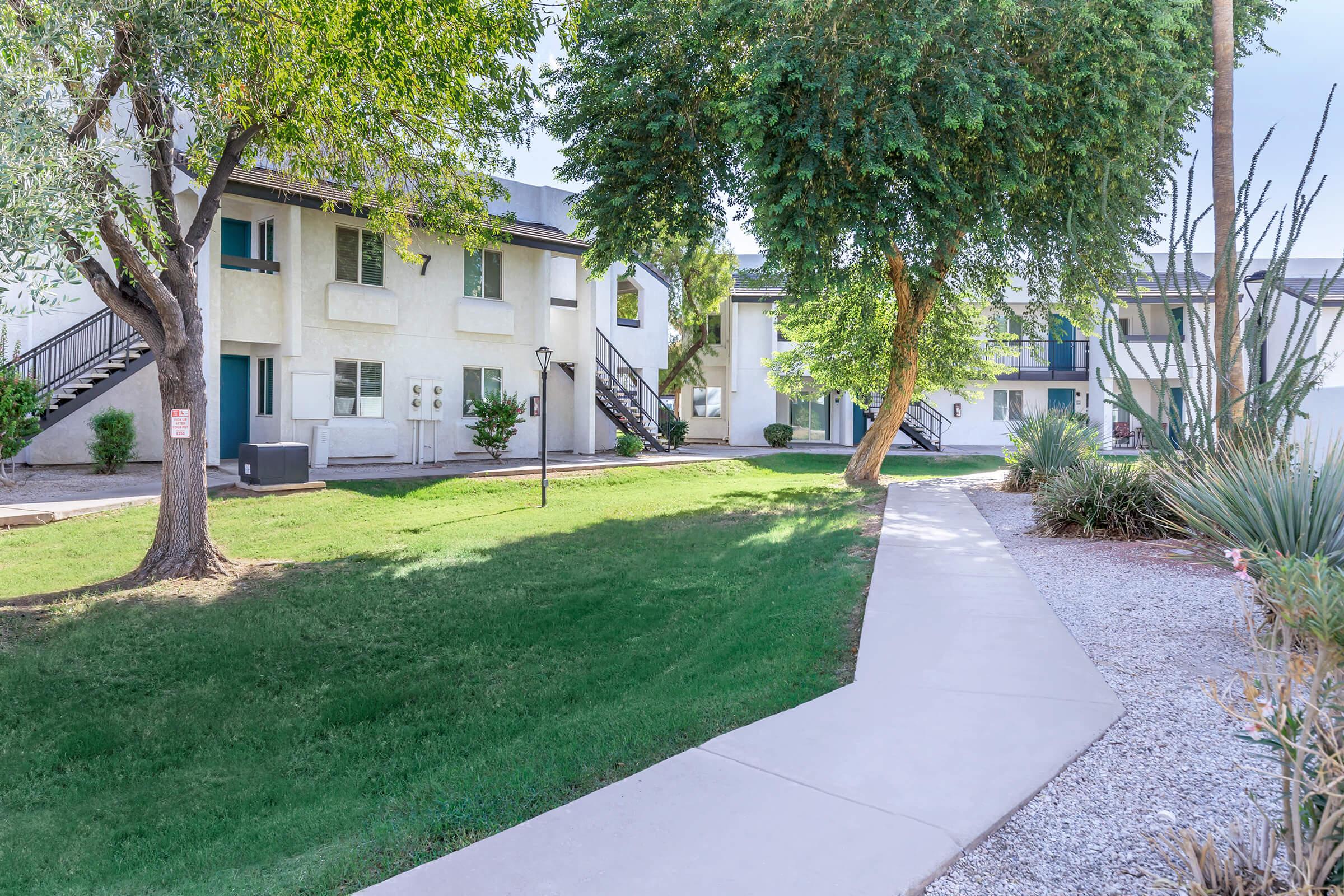 A landscaped courtyard featuring green grass and trees, with paths leading between two-story residential buildings. The scene includes a walkway, grassy areas, and lamp posts, creating a serene outdoor space in an apartment complex.