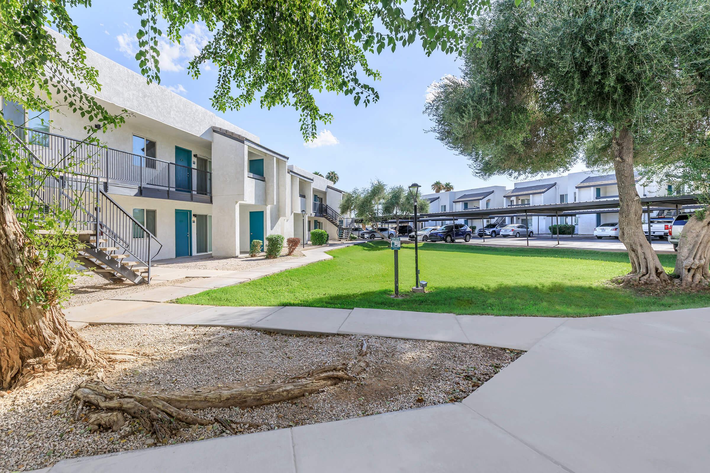A view of a residential complex with two-story buildings and green landscaping. The scene features a grassy area, a tree, and a walkway. Apartments are visible in the background, with parking spaces for vehicles. The sky is clear and sunny, creating a bright and inviting atmosphere.