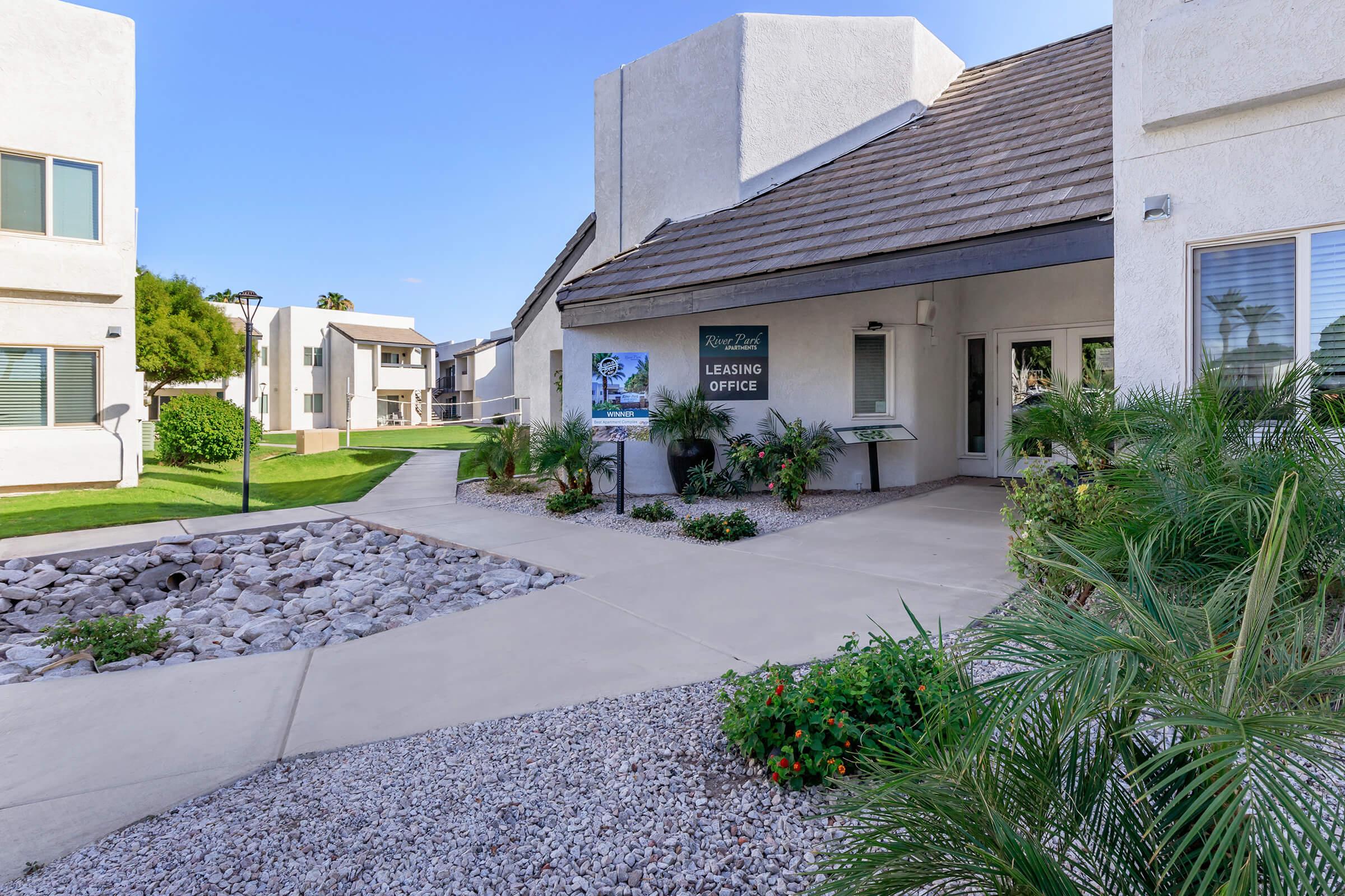 A leasing office at a modern apartment complex, surrounded by neatly landscaped greenery. The entrance features a sign indicating the office, and there are smooth pathways lined with rocks and plants, creating a welcoming atmosphere. Clear blue skies are visible in the background.