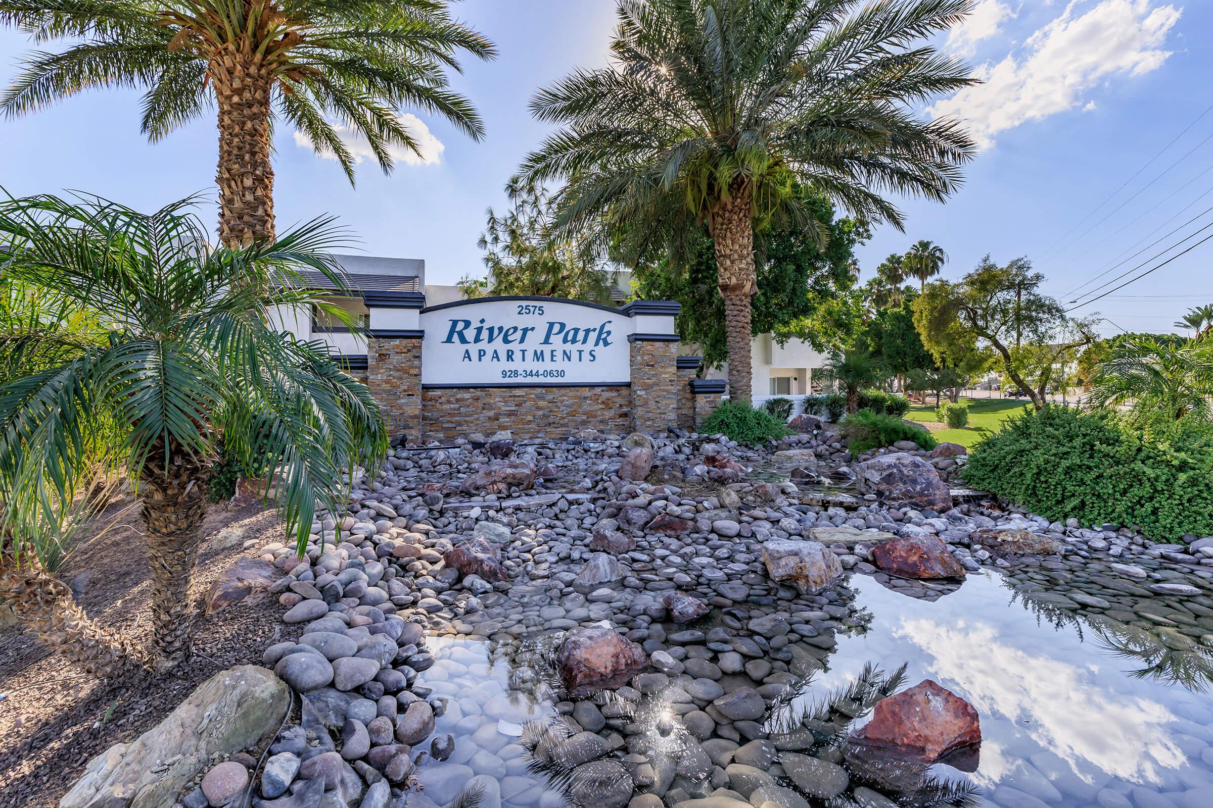 Sign for River Park Apartments surrounded by lush greenery, palm trees, and decorative rocks. A small pond with reflections adds to the serene atmosphere. The sign includes the address "2975" and a contact number. The scene captures a welcoming entrance to the apartment complex.