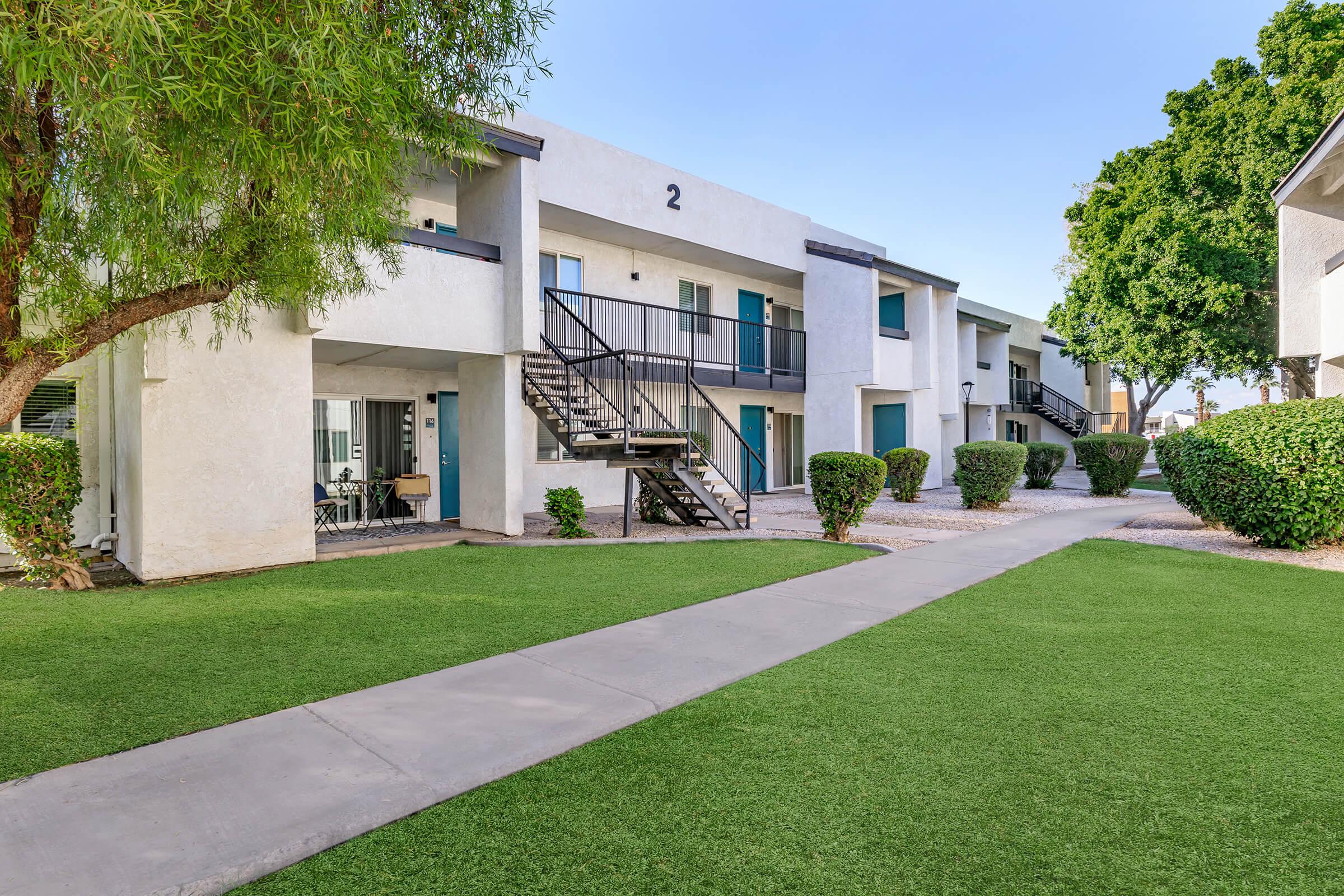 Two-story apartment buildings with a well-maintained lawn and pathway. The buildings feature a white exterior with blue doors and small balconies. Lush greenery surrounds the area, including trees and hedges, creating a welcoming environment. Clear blue sky above adds to the pleasant appearance.