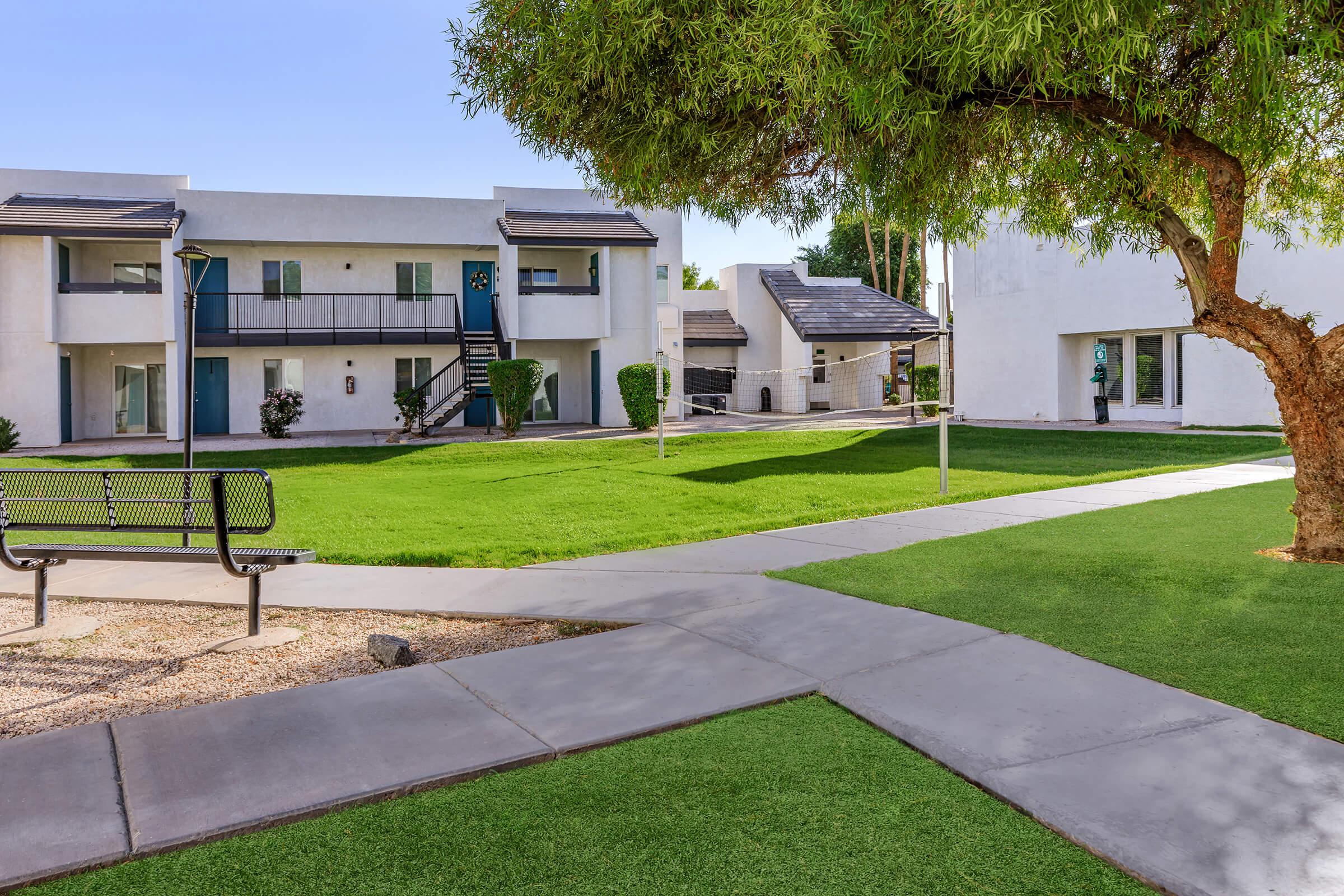 A landscaped courtyard with green grass and a tree, surrounded by white apartment buildings. A black bench is visible on a paved walkway that leads through the area. The setting appears well-maintained and inviting.