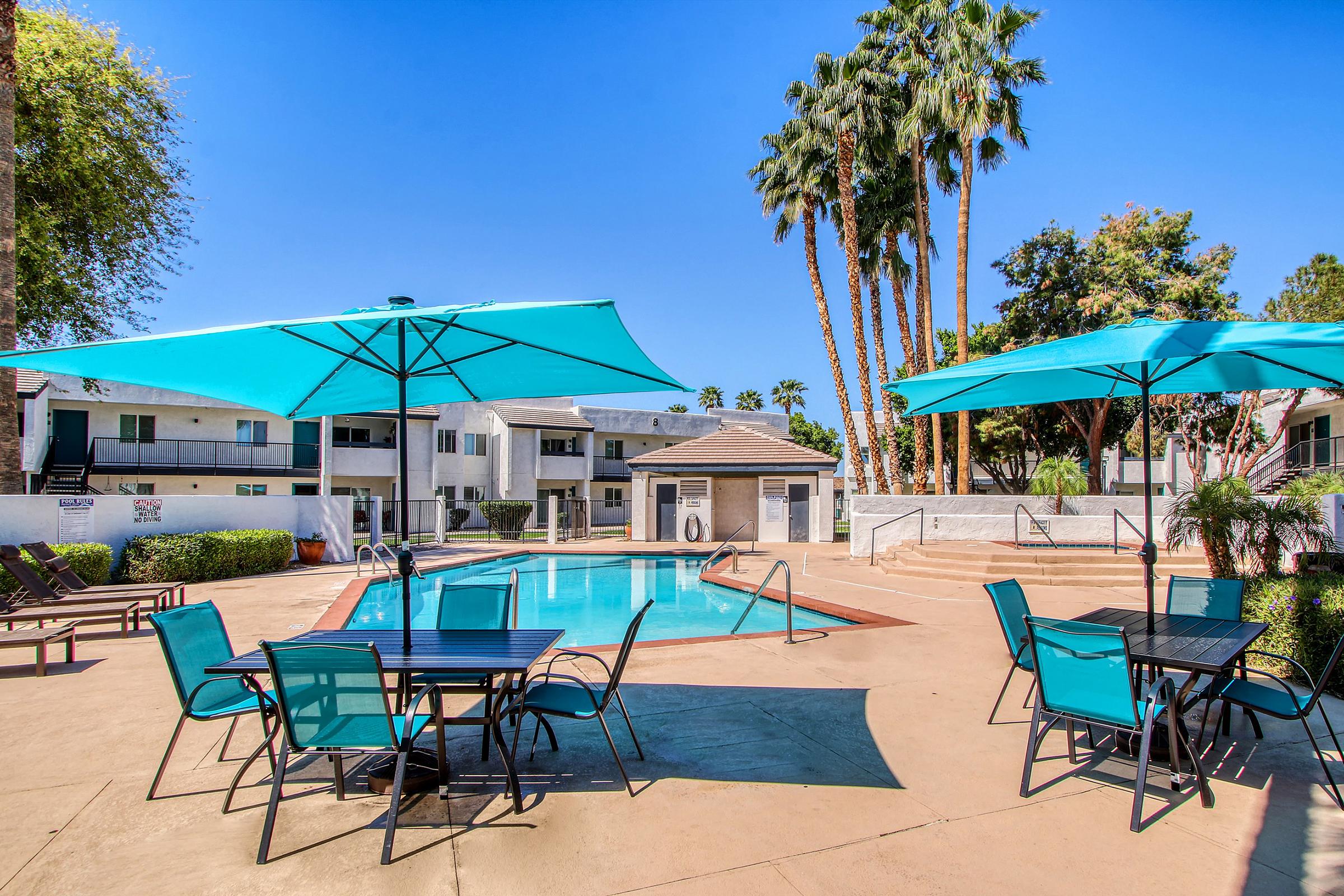 A sunny outdoor pool area featuring bright blue umbrellas and patio furniture. Palm trees surround the space, and nearby buildings are visible in the background. The clear blue sky adds to the inviting atmosphere of the pool area.