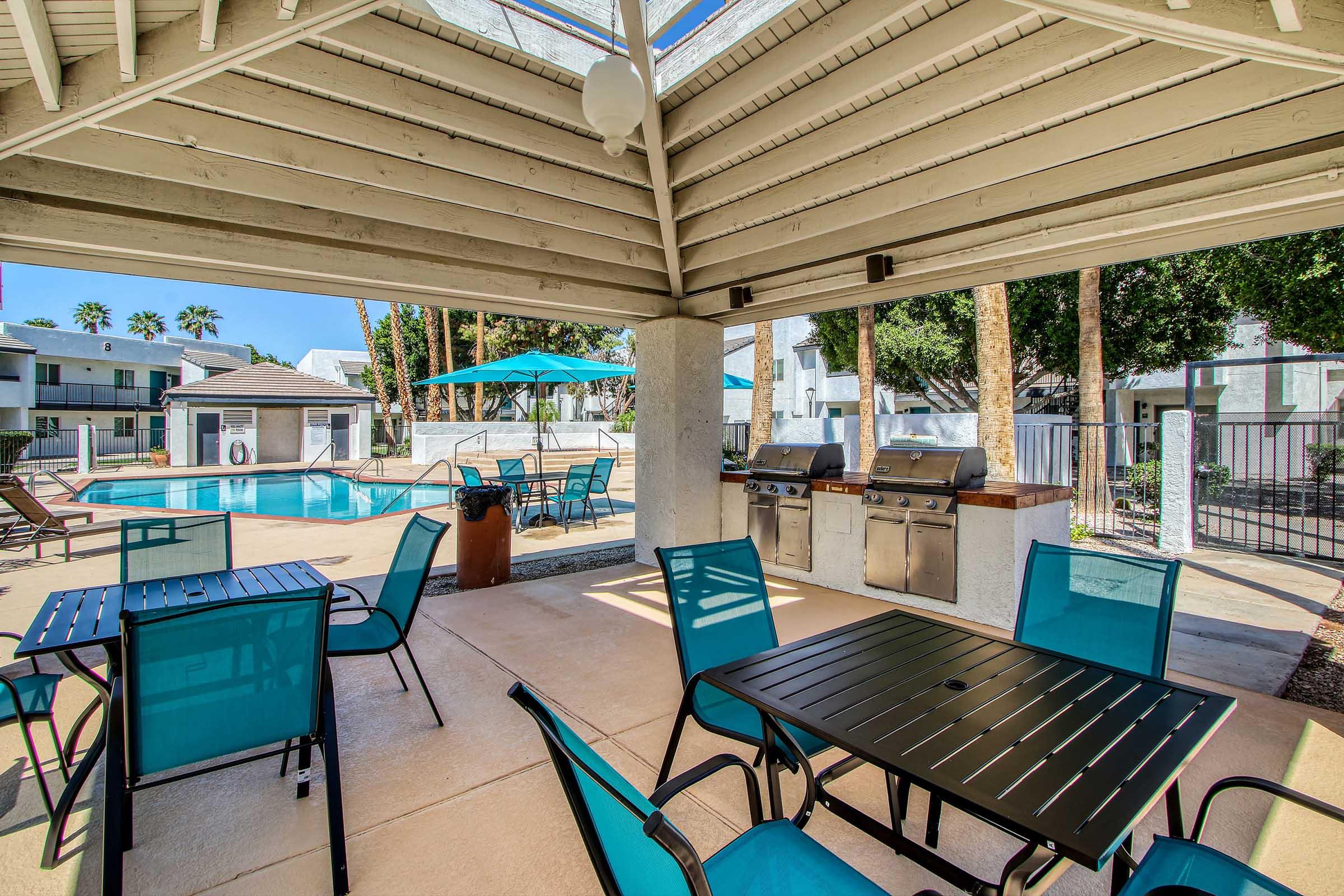 A shaded outdoor area with blue tables and chairs overlooking a swimming pool. There are barbecue grills and umbrellas for shade. Surrounding palm trees and well-maintained landscaping create a relaxing atmosphere in a residential community setting.