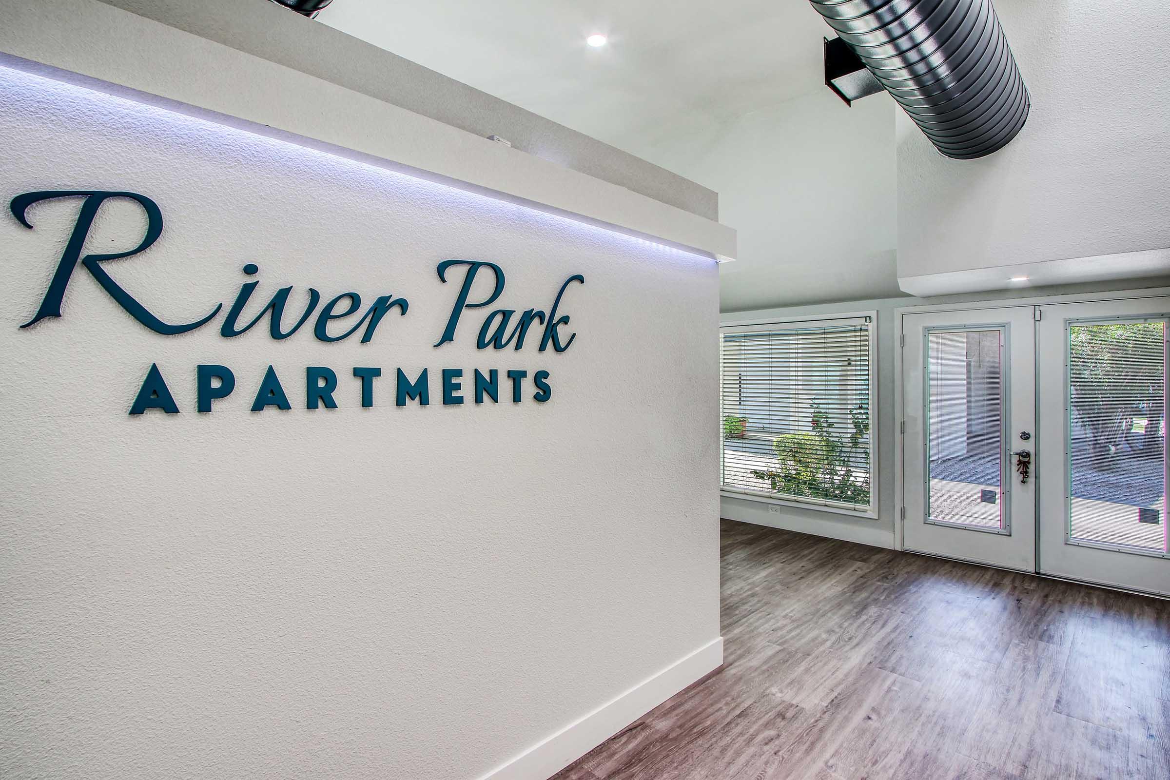 Interior view of the River Park Apartments entrance, featuring a modern design. The sign "River Park Apartments" is prominently displayed on a white wall, with natural light streaming in through large windows. The space has wood-like flooring and a contemporary feel.