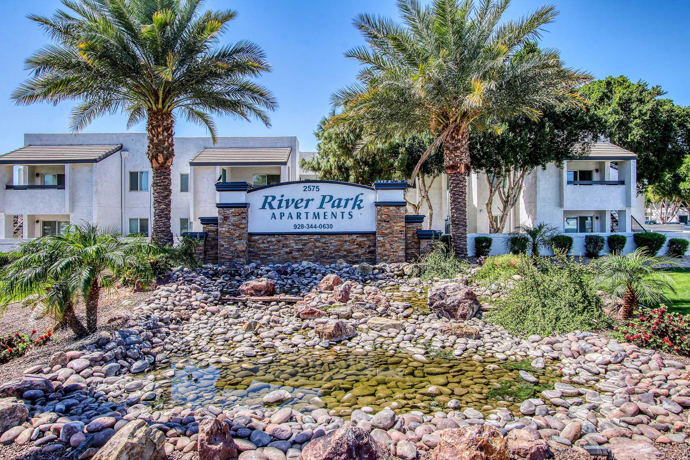 Sign for River Park Apartments at 2575 with a landscaped area featuring palm trees, rocks, and a small water feature in front of a white apartment building. The scene represents a welcoming residential environment.