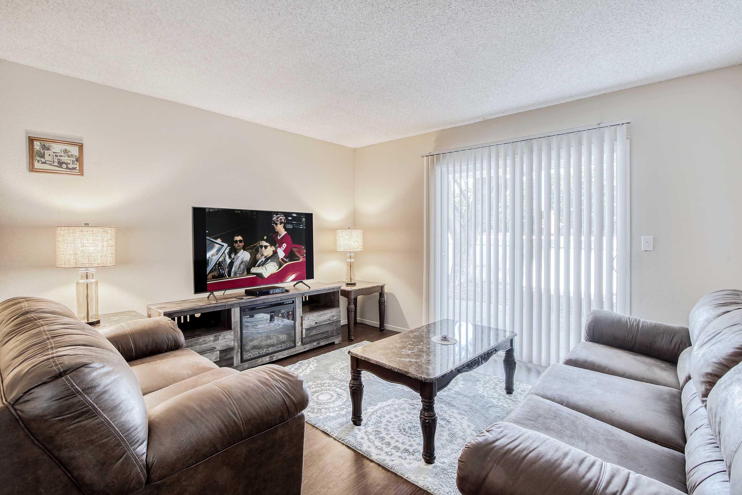 A cozy living room featuring two brown leather sofas, a wooden coffee table, and a flat-screen TV displaying a scene. There are two decorative lamps on either side of the TV stand, and large sliding glass doors with vertical blinds allow natural light to enter the space. The room has neutral wall colors and a soft area rug.
