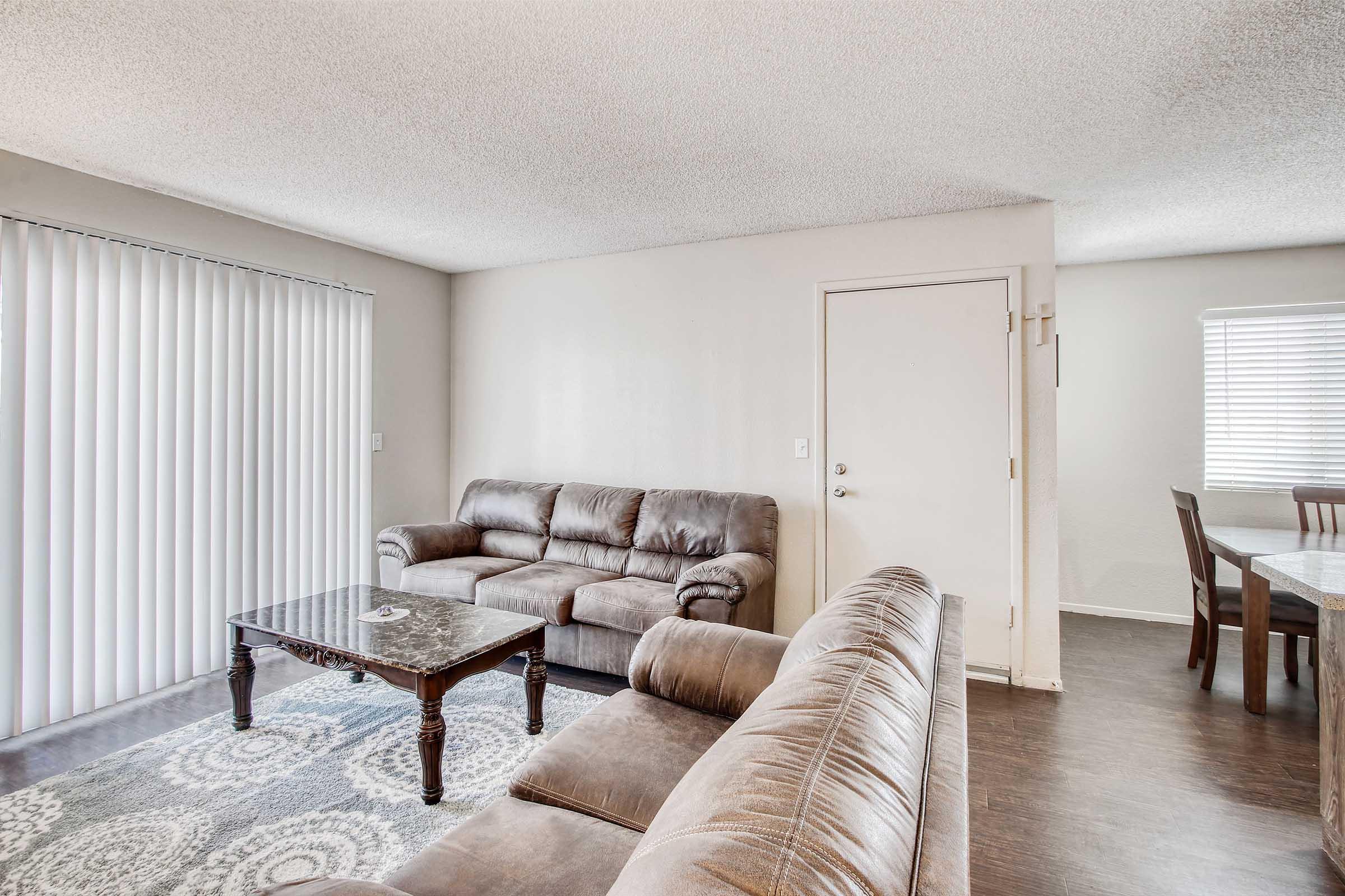 A cozy living room featuring a light brown leather sofa set, a dark wood coffee table, and a dining area with a wooden table. Natural light enters through vertical blinds, enhancing the neutral color scheme of the walls and flooring. A door leads outside, contributing to the inviting atmosphere.