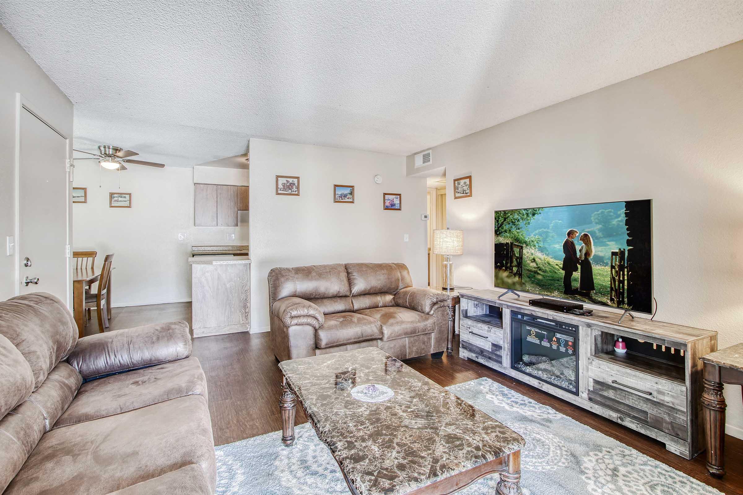Cozy living room featuring brown leather sofas, a coffee table, and a television displaying a romantic scene. Light-colored walls and wooden furniture create a warm atmosphere. The background includes a dining area and kitchen, enhancing the inviting ambiance of the space.