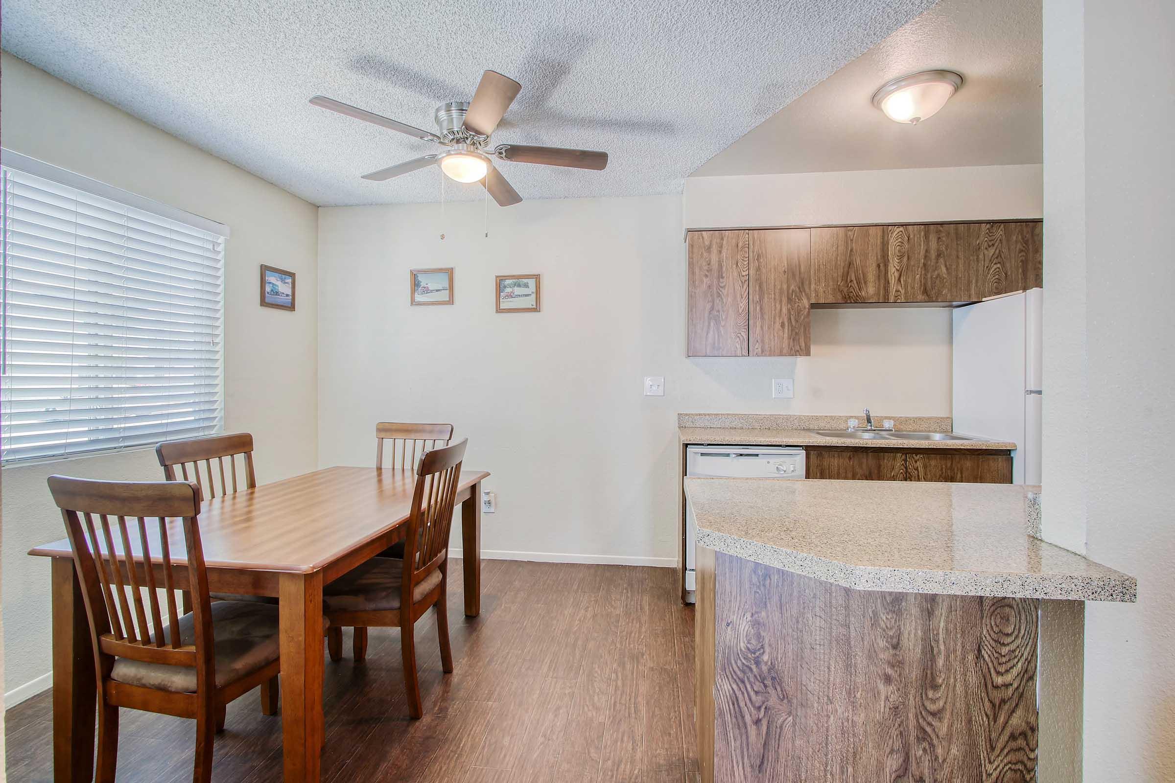 A small kitchen and dining area featuring a wooden table with four chairs, a ceiling fan, and light-colored walls. There's a countertop with cabinets and a sink, along with kitchen appliances partially visible. Natural light enters through a window adorned with blinds and framed pictures on the wall.