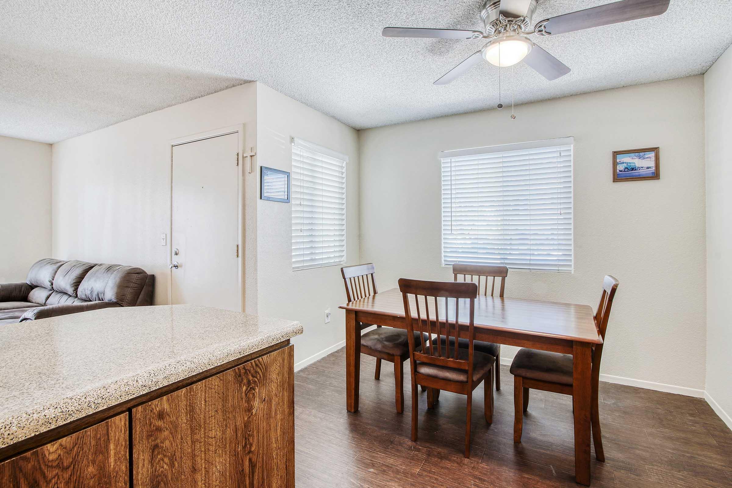 A dining area with a wooden table and four chairs, featuring a ceiling fan and windows with blinds. A living area is visible in the background, with a couch and a door leading outside. The walls are light-colored, and there is a framed picture on the wall near the windows.