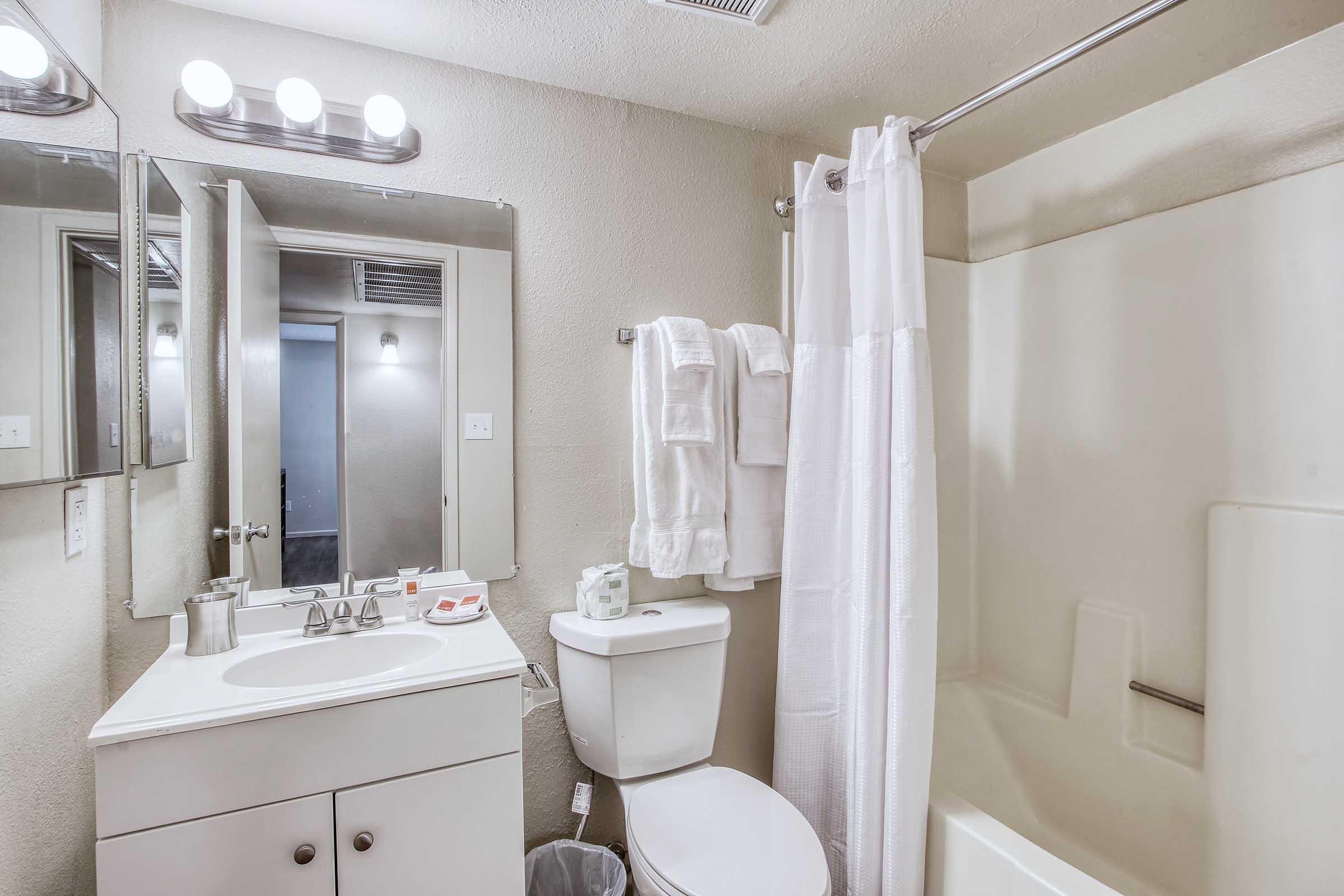 A bathroom featuring a bathtub with a shower curtain, a white toilet, and a sink with a mirror above it. White towels are neatly hung on a rack. The walls are painted in a light color, and there’s a small trash can next to the toilet. Soft lighting from overhead fixtures enhances the clean and tidy appearance.