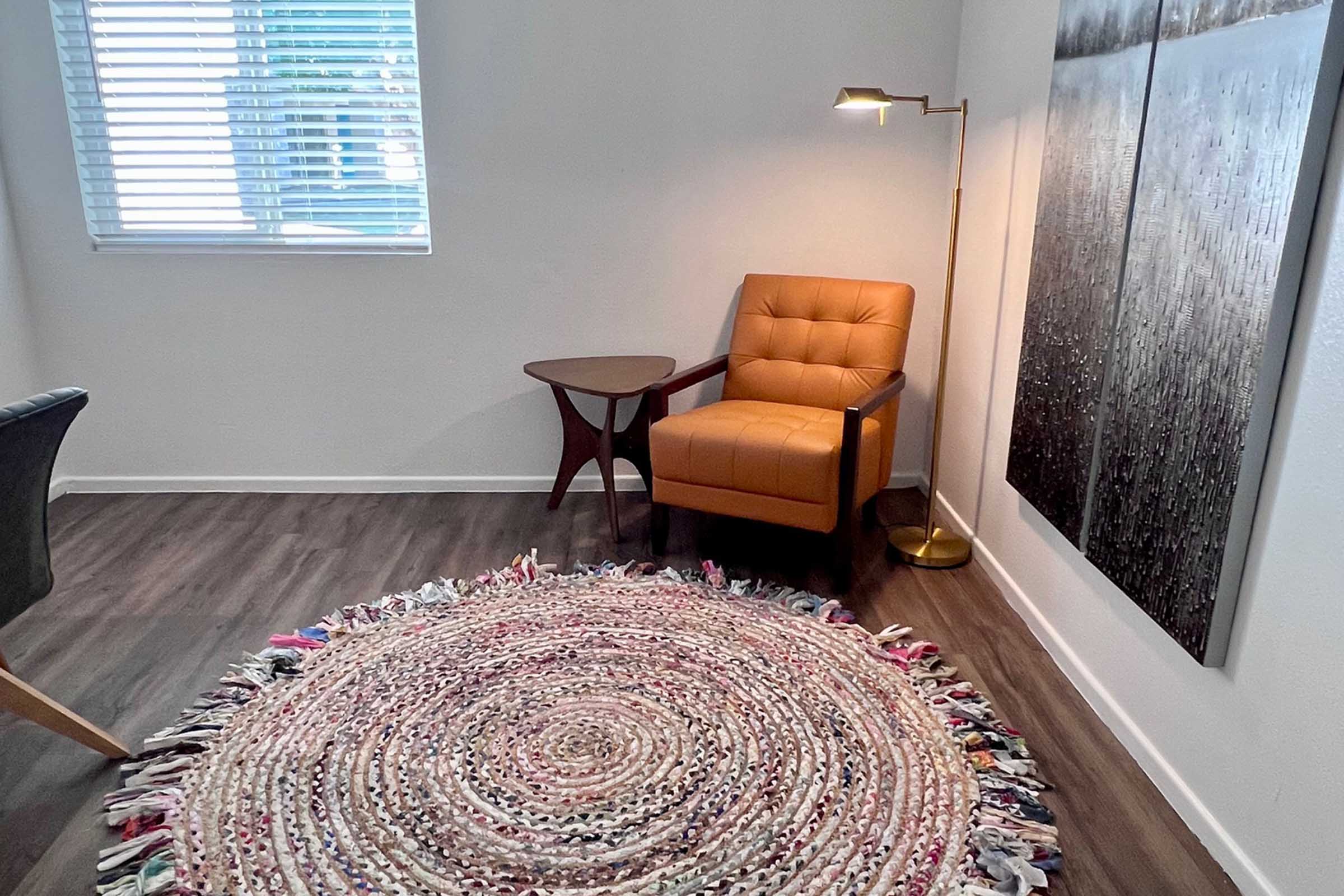 A cozy corner of a room featuring a brown leather armchair beside a small wooden side table. A stylish standing lamp illuminates the space. The floor is adorned with a round, multicolored woven rug, and the walls display two large abstract art pieces, adding a modern touch to the serene ambiance.