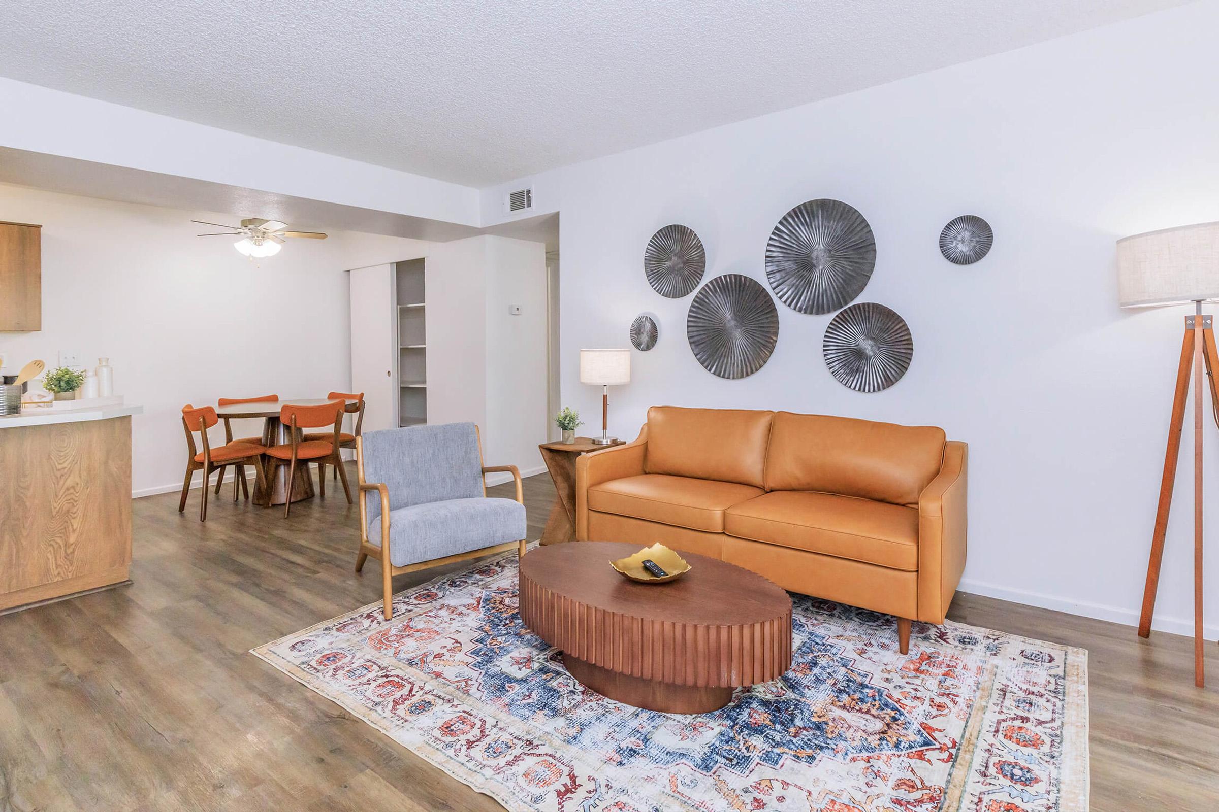 A modern living room featuring a stylish brown leather sofa, a round wooden coffee table with a decorative bowl, and a light gray chair. The walls have circular metal art pieces, and a floor lamp stands beside the sofa. In the background, a dining area with a wooden table and chairs is visible, along with a ceiling fan.