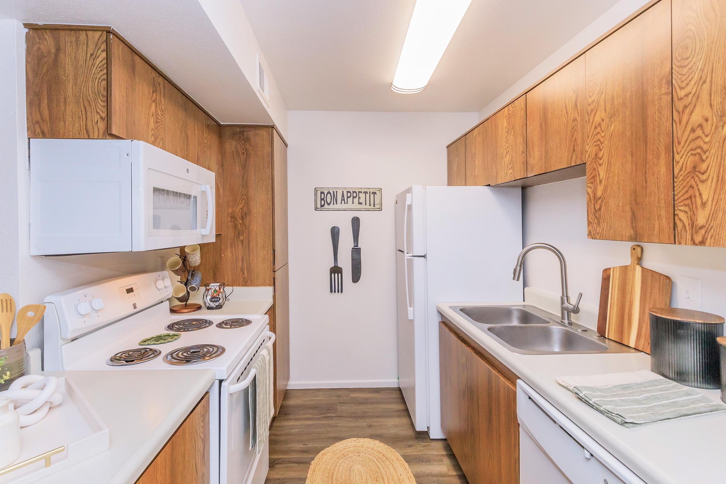 A cozy kitchen featuring wooden cabinets, white appliances, and a minimalist design. The space includes a stove, microwave, sink, and a cutting board on the counter, along with utensils displayed on the wall, creating an inviting cooking environment.