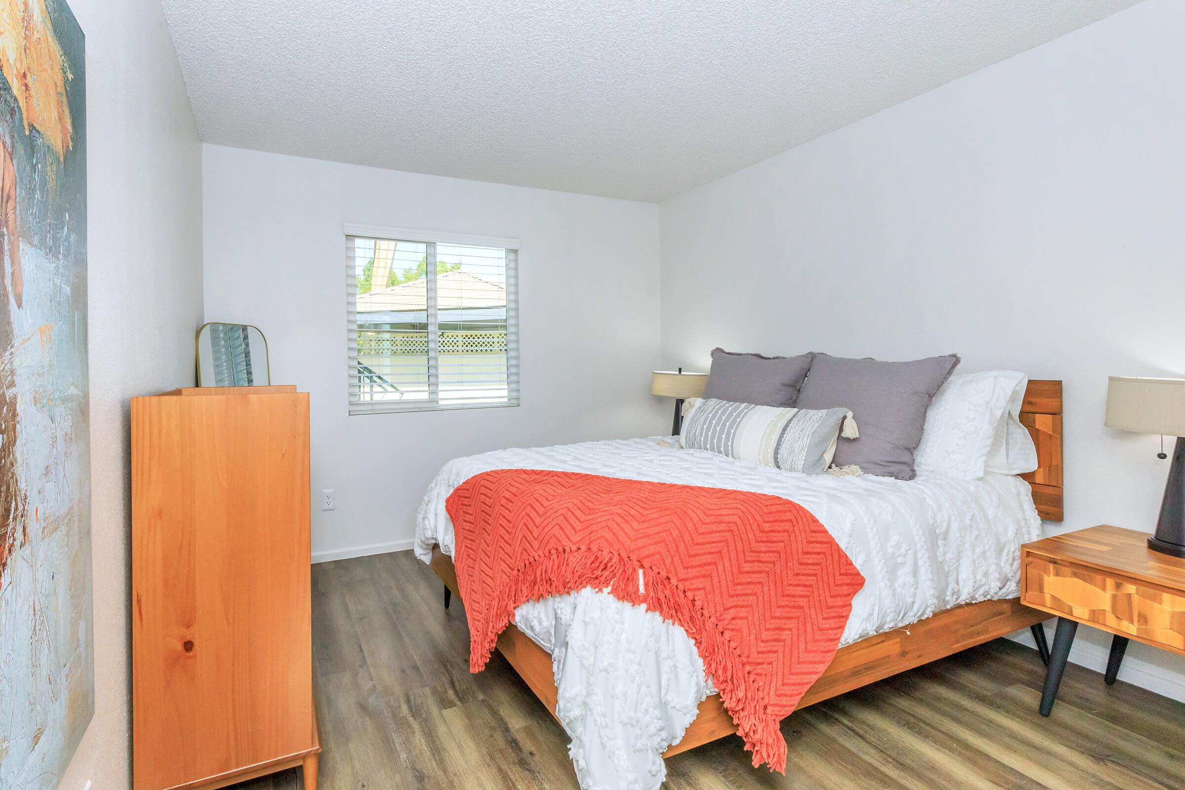 A cozy bedroom featuring a wooden bed with a white comforter and an orange blanket. There are two bedside lamps on wooden nightstands, a wooden dresser, and a large abstract painting on the wall. A window allows natural light to enter, and the flooring is a light wood finish.