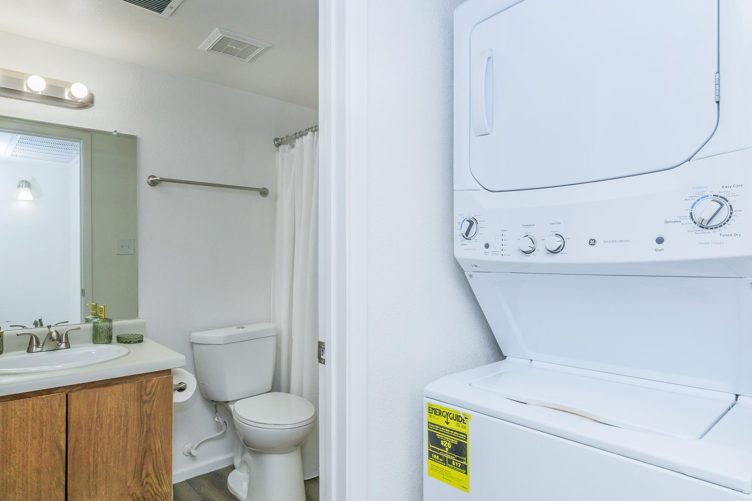 A compact laundry area featuring a stacked washer and dryer next to a bathroom. The bathroom includes a white toilet, a sink with wooden cabinetry, and a mirror. The walls are painted white, and there is a shower curtain partially visible. The space is well-lit and designed for utility and convenience.