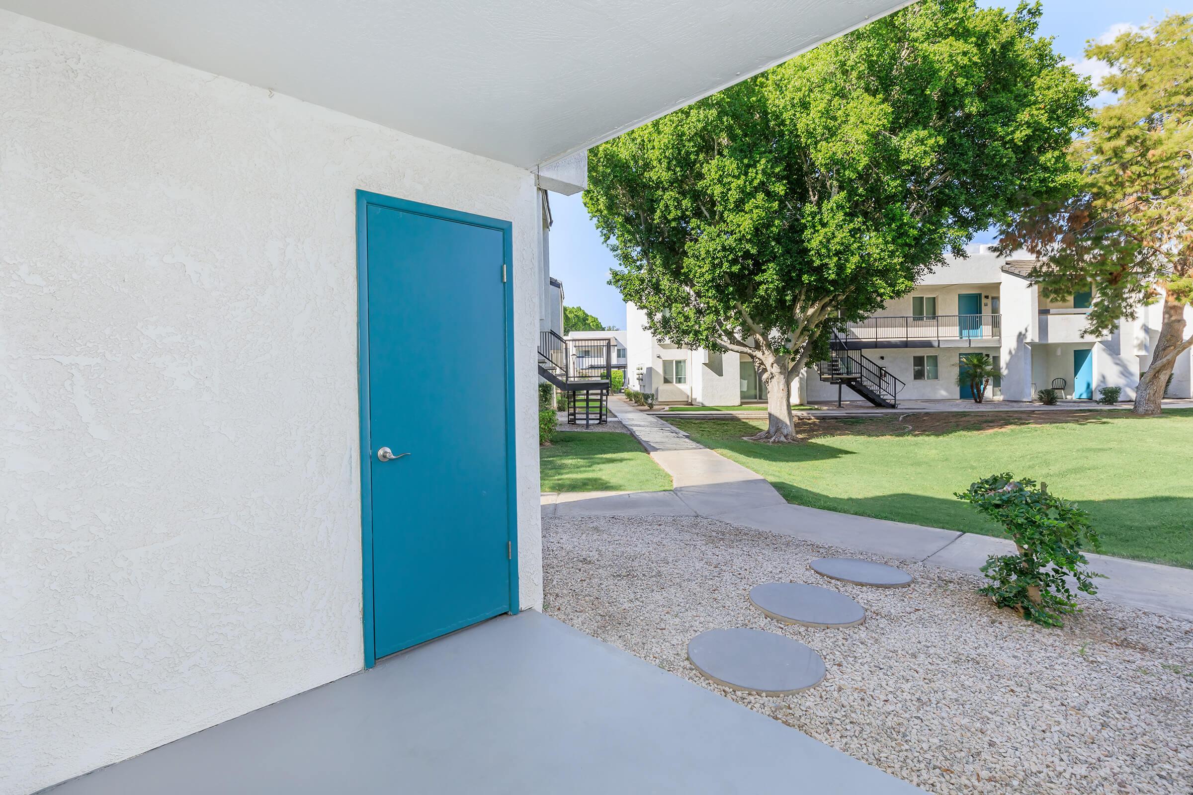 A view from a doorway showing a blue door on a white wall, leading to a landscaped area with green grass, a pathway of stepping stones, and trees. In the background, there are apartment buildings with stairs and balconies visible. Bright and inviting outdoor setting.