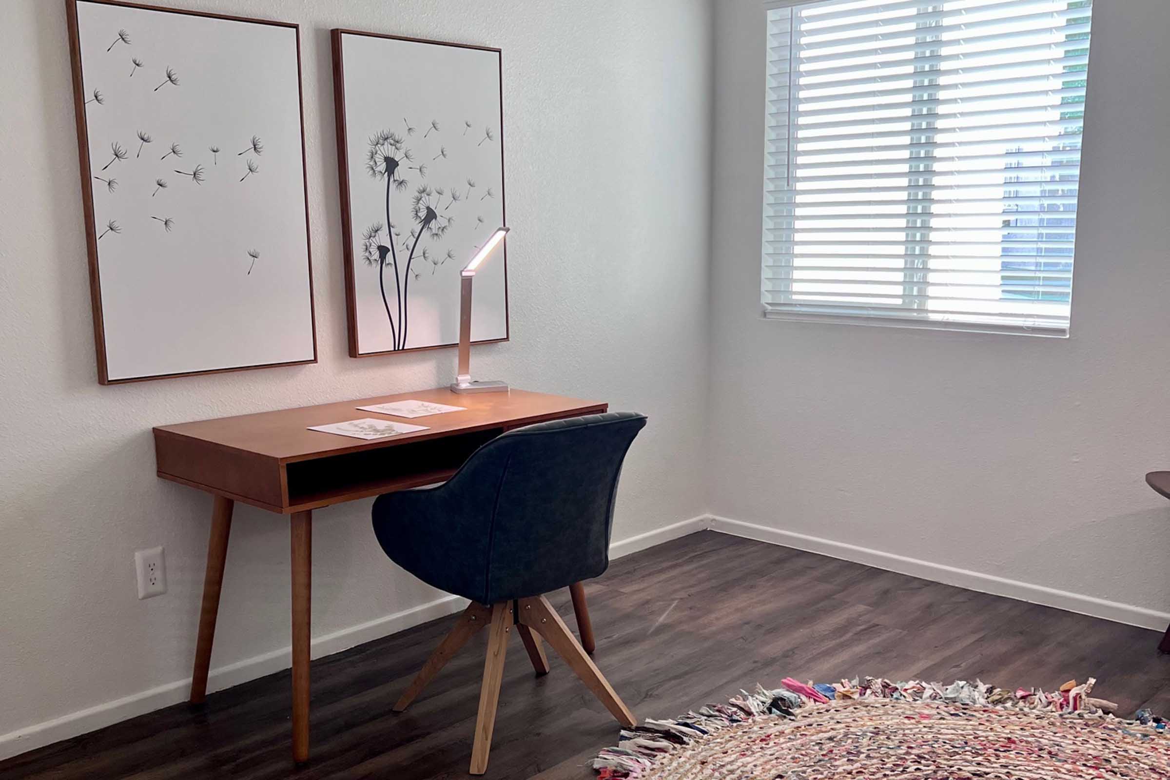 A minimalist workspace featuring a wooden desk with a lamp, a blue upholstered chair, and decorative art prints on the wall. A circular, colorful rug is on the floor, and a window with blinds allows natural light to illuminate the room.