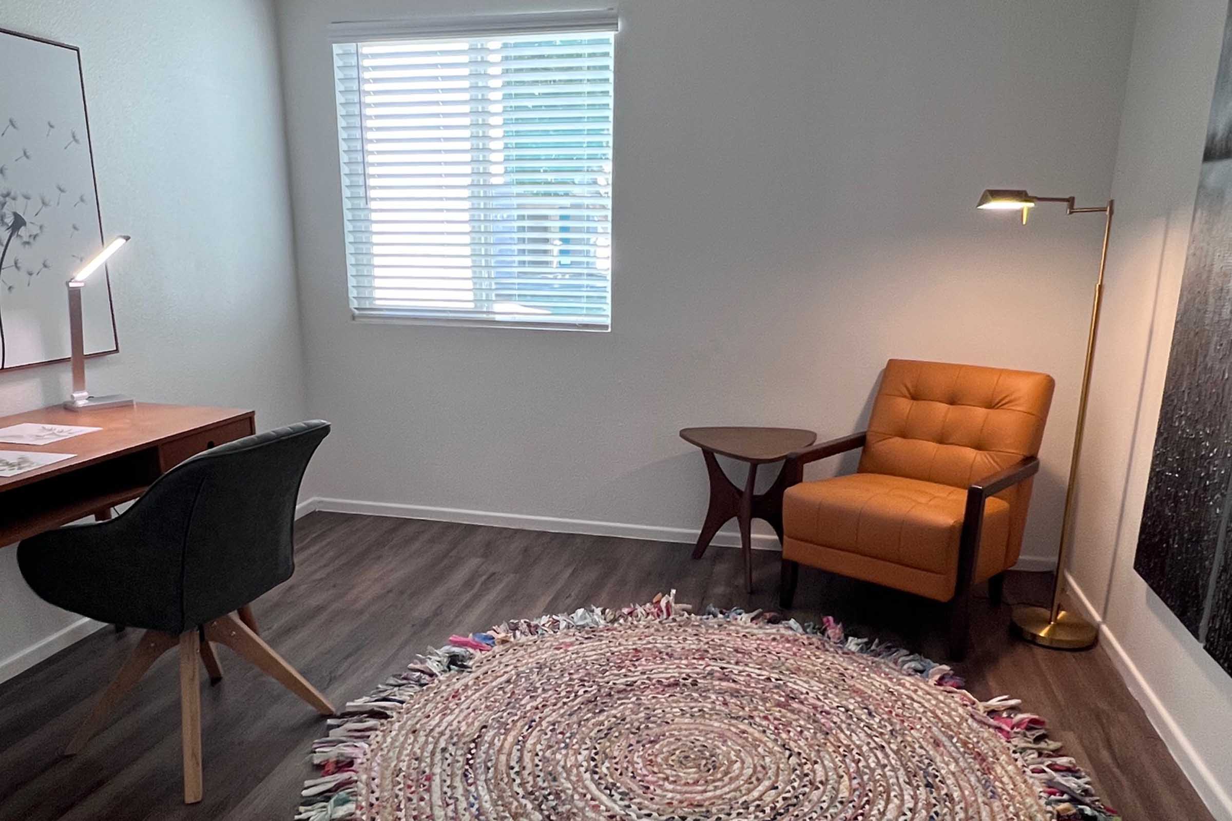 A cozy interior space featuring a wooden desk with papers, a black chair, an orange armchair, a round braided rug, and a floor lamp. Natural light streams in through a window with blinds, creating a warm and inviting atmosphere.