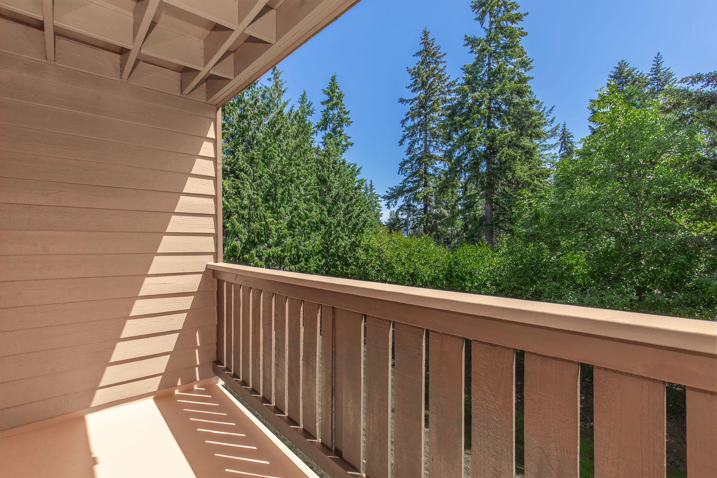 A wooden balcony with a light brown railing, overlooking a dense green forest of tall trees under a clear blue sky. Sunlight casts shadows on the balcony floor, creating a warm and inviting atmosphere.