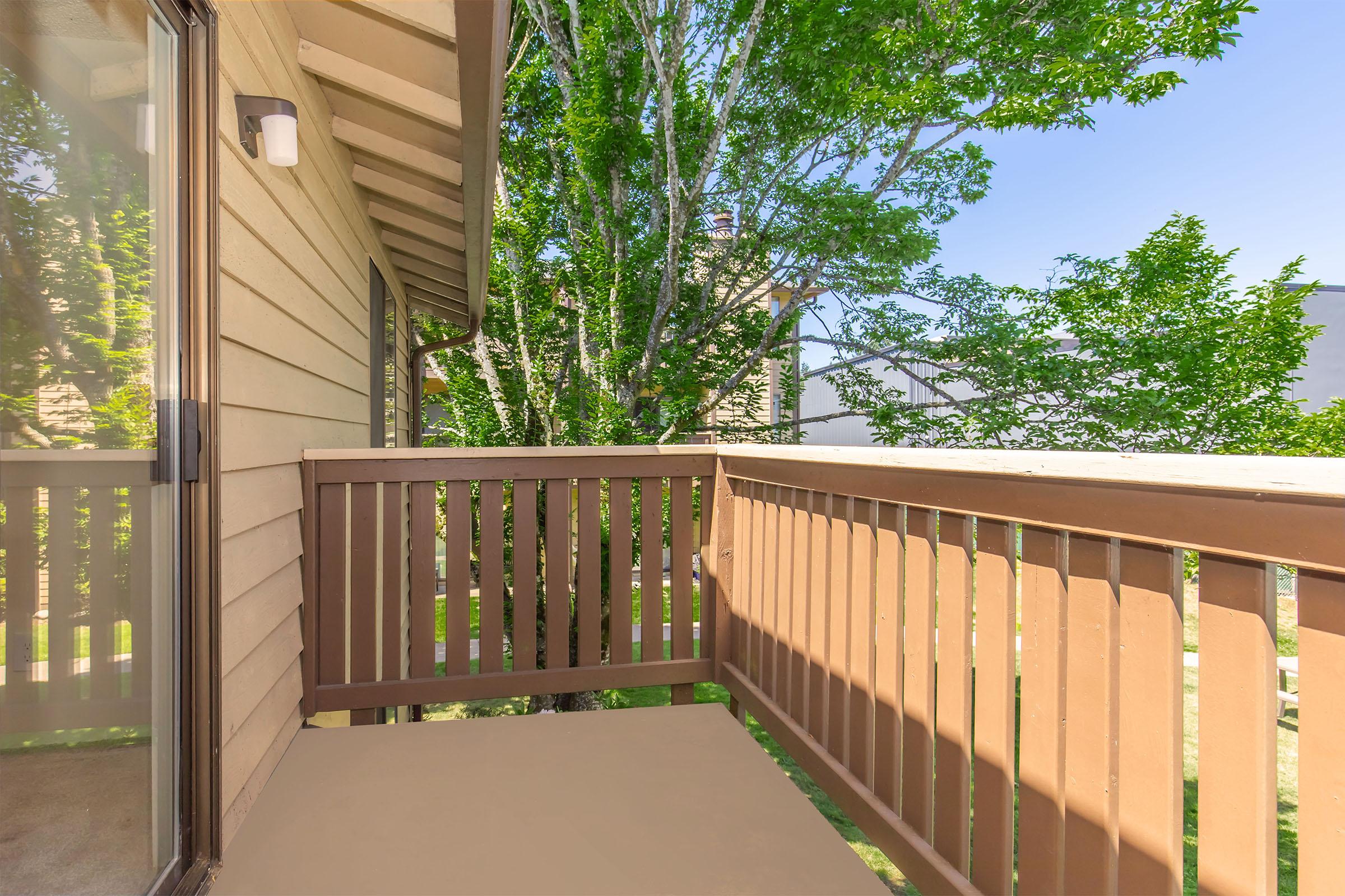 A corner balcony with a wooden railing, surrounded by greenery. The sunlight illuminates the space, showcasing the wooden floor and the building's exterior. Trees and blue sky are visible in the background, creating a serene outdoor atmosphere.
