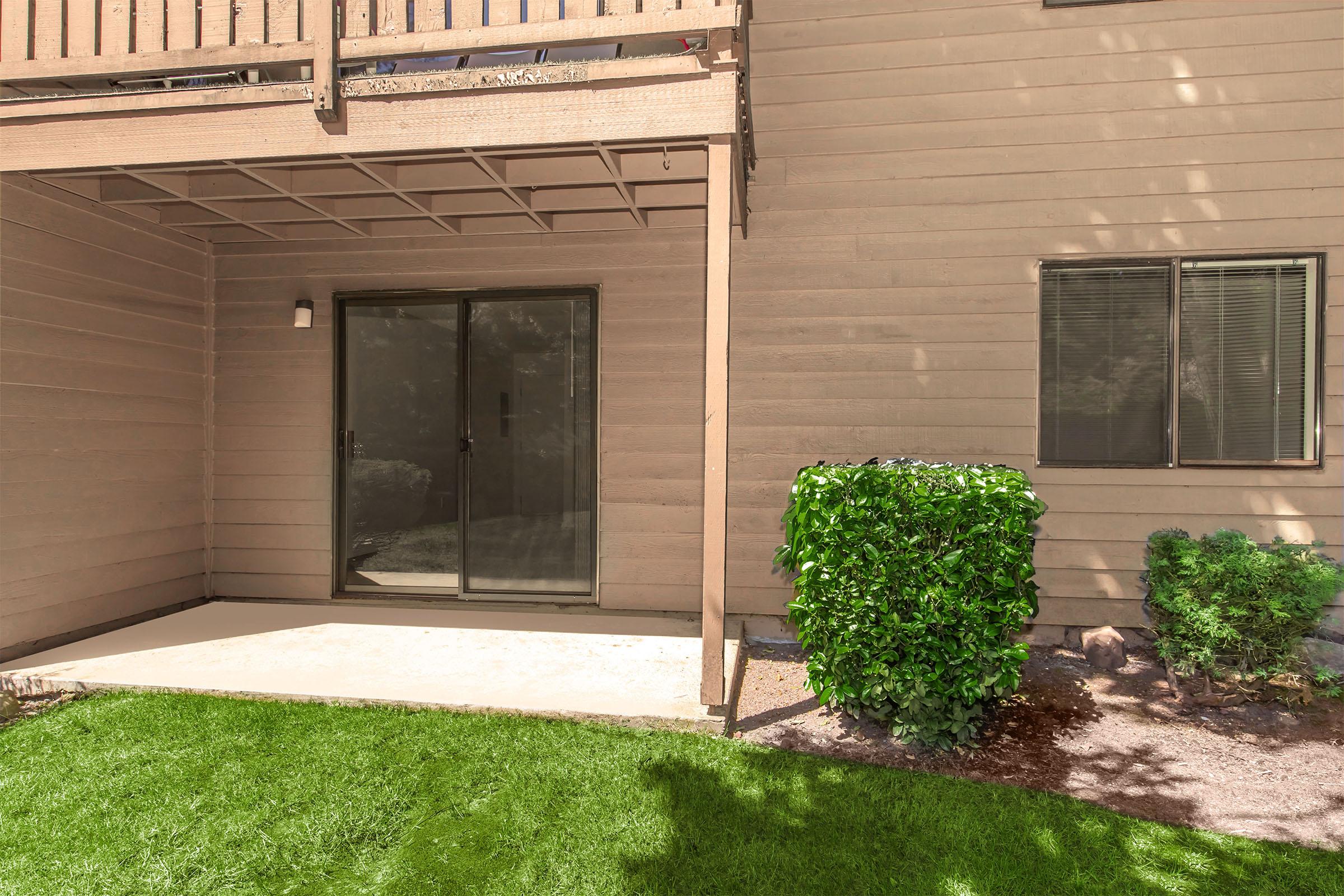 A ground-level patio area with sliding glass doors, surrounded by grass and small shrubs. The exterior of the building is light brown with a wooden balcony above. Two windows are visible, framed by the beige wall, with one window partially obscured by greenery.