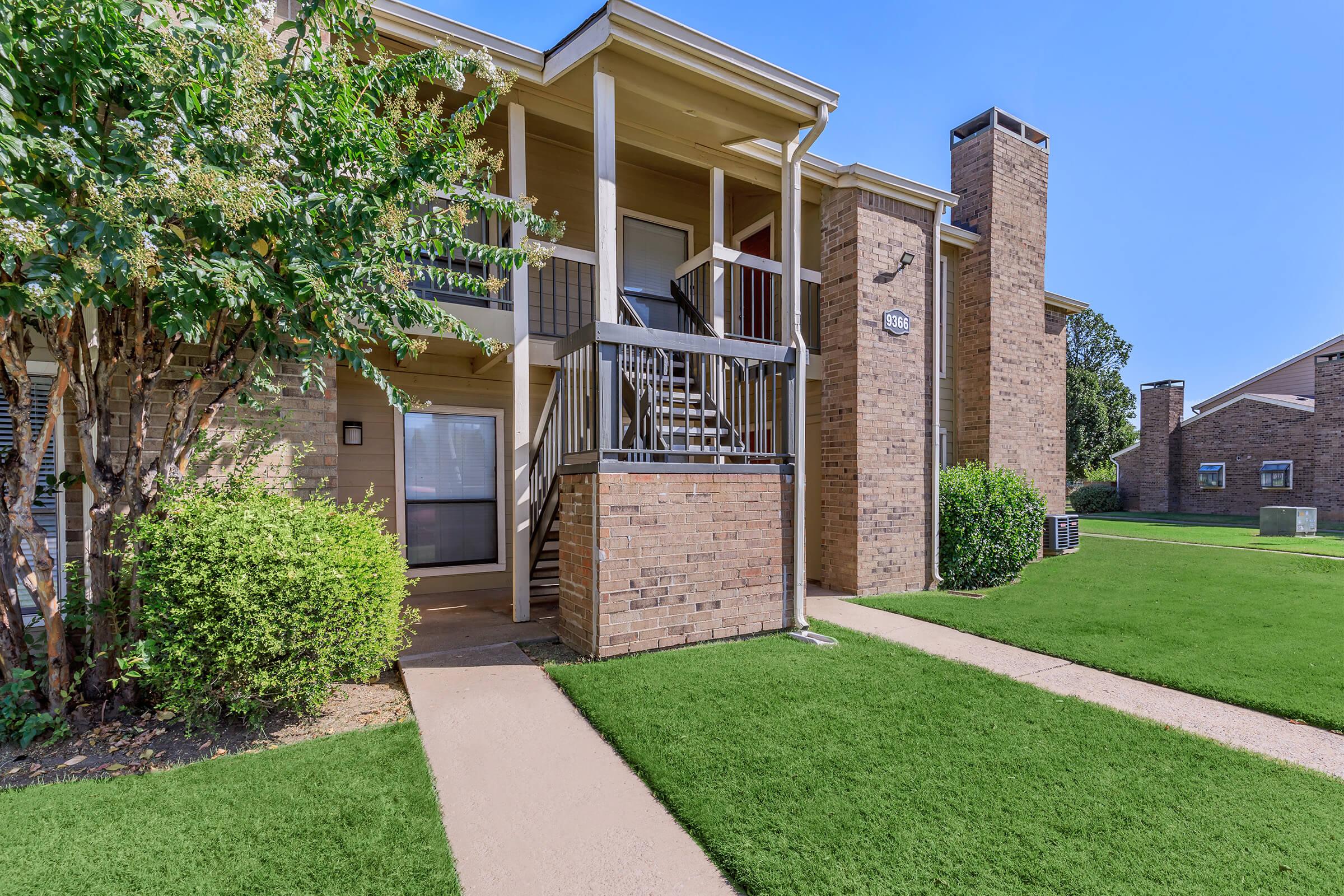 Exterior view of a multi-story apartment building featuring a brick facade, green grass, and landscaped bushes. The entrance has a staircase leading up to a balcony, with clear blue skies in the background. Pathways are visible leading to the building and surrounding structures.