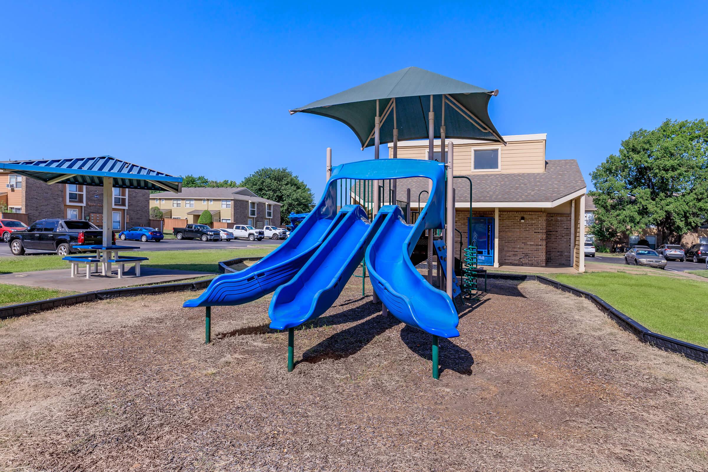 A children's playground featuring two blue slides under a shaded canopy, surrounded by grassy areas. In the background, there are several apartment buildings and a picnic area with tables. The sky is clear and blue, creating a bright, inviting atmosphere.