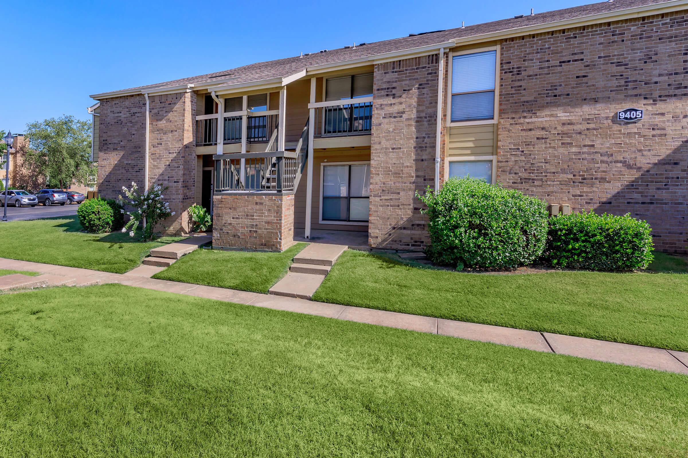 Brick apartment building with two levels, featuring balconies and a well-manicured lawn. There are concrete walkways leading to the entrance and green shrubbery along the sides. The sky is clear and blue, and the scene suggests a sunny day.