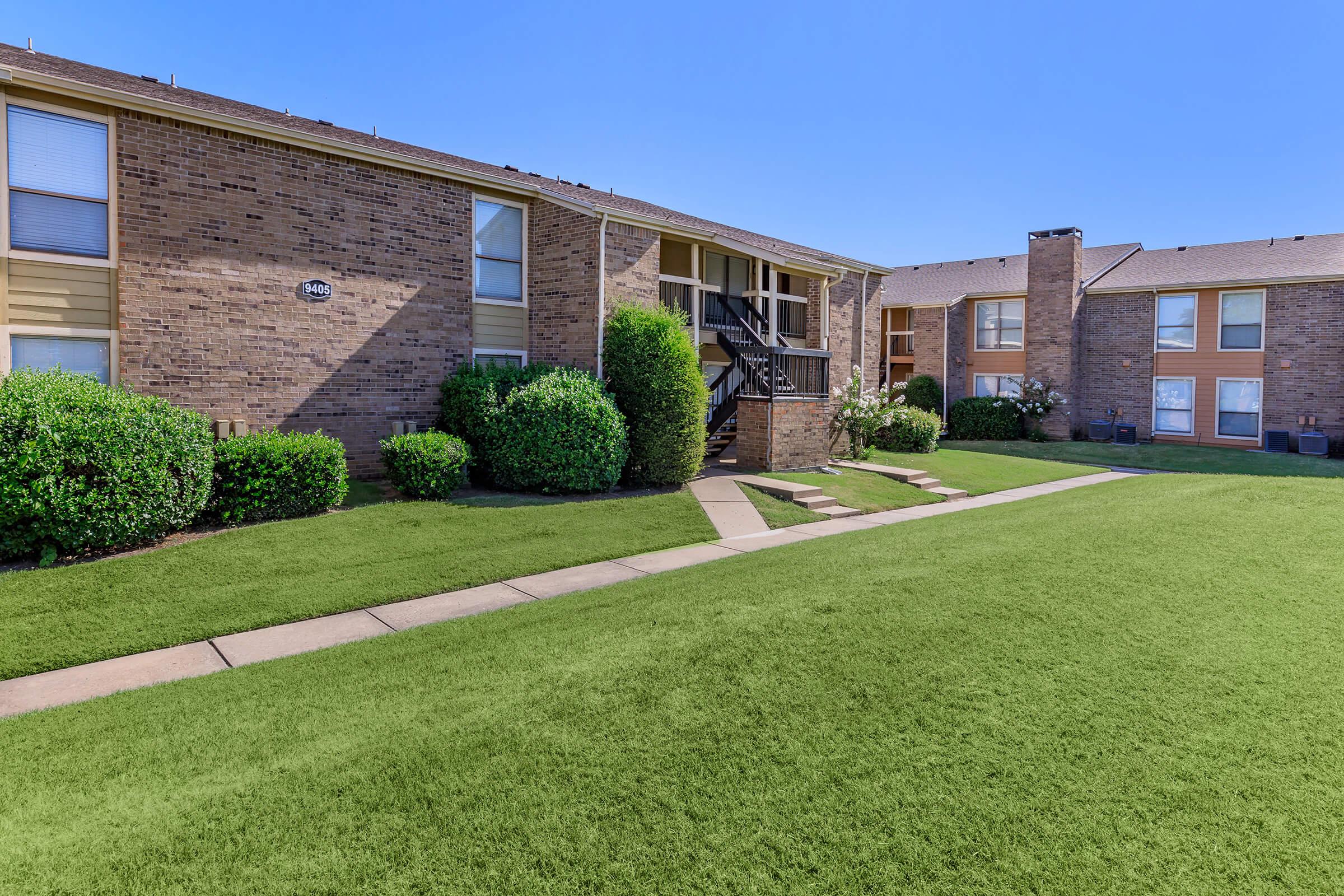 Brick apartment complex with a green lawn in front, featuring two-story buildings and well-maintained shrubs. Clear blue sky above, with a path leading to the entrance.