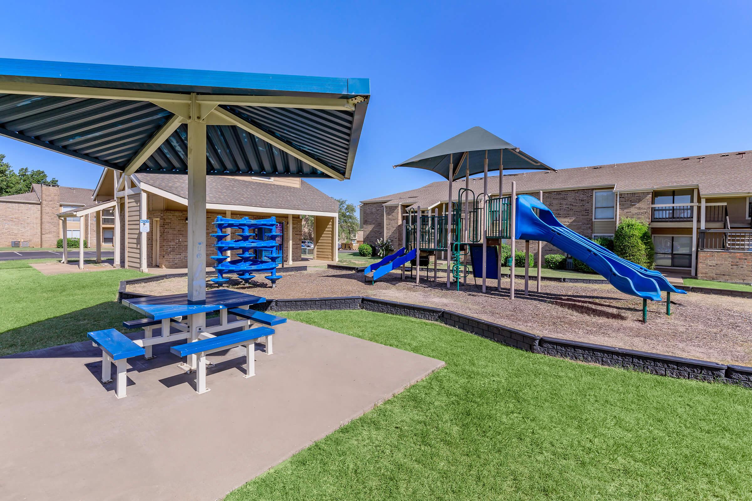 A playground area featuring blue slides and a climbing structure, surrounded by grassy areas. There is a covered picnic table with blue benches nearby, and residential buildings in the background under a clear blue sky.