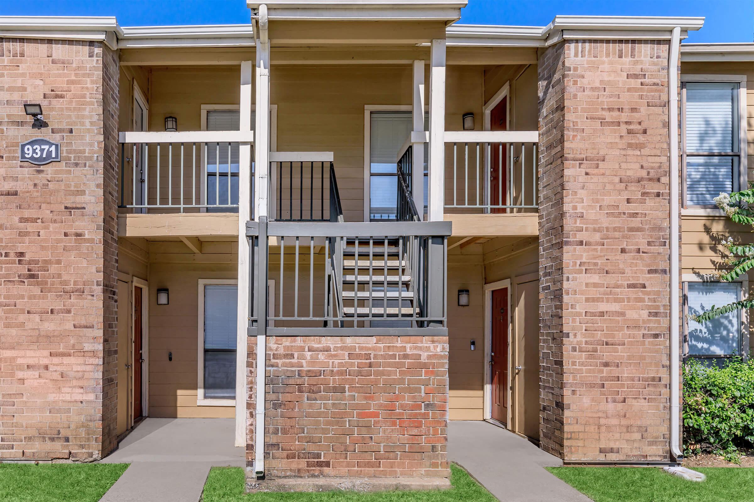 Exterior view of a two-story apartment building featuring brick and wooden elements. The image shows two sets of stairs leading to separate entryways with beige walls and white shutters. A small grass area is visible in front, and the building is well-lit by natural sunlight.