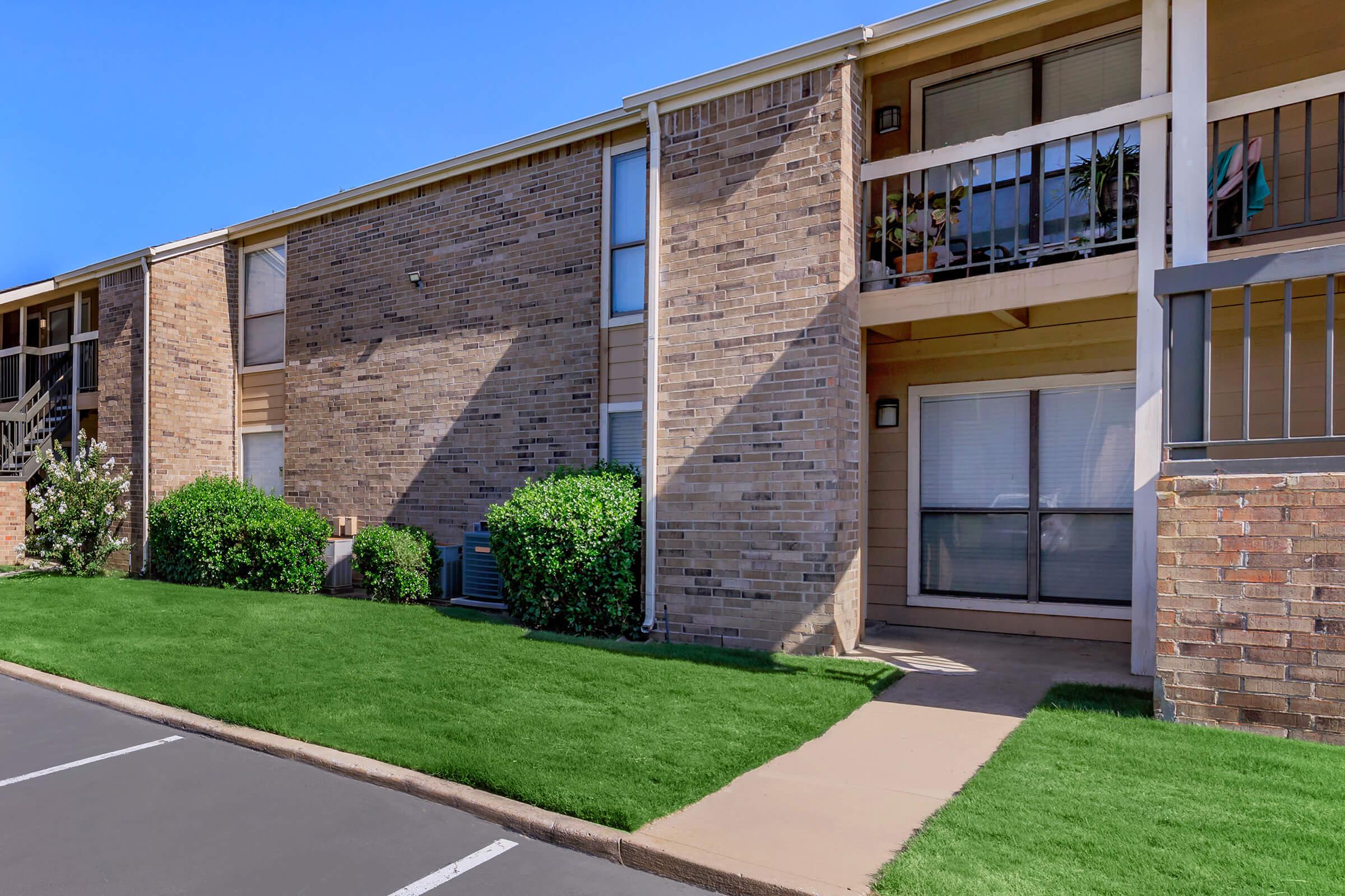 A view of a two-story brick apartment building with a well-maintained lawn. The building features balconies on the upper level, surrounded by shrubs. A paved pathway leads to the entrance, and parking spaces are visible in the foreground. Clear blue sky above enhances the scene.