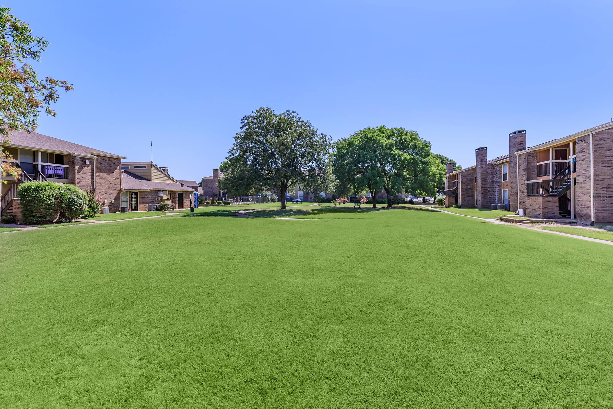 A sunny landscape featuring a lush green lawn surrounded by two sets of apartment buildings. In the center, there are two large trees providing shade. The clear blue sky adds to the bright and inviting atmosphere of the area.