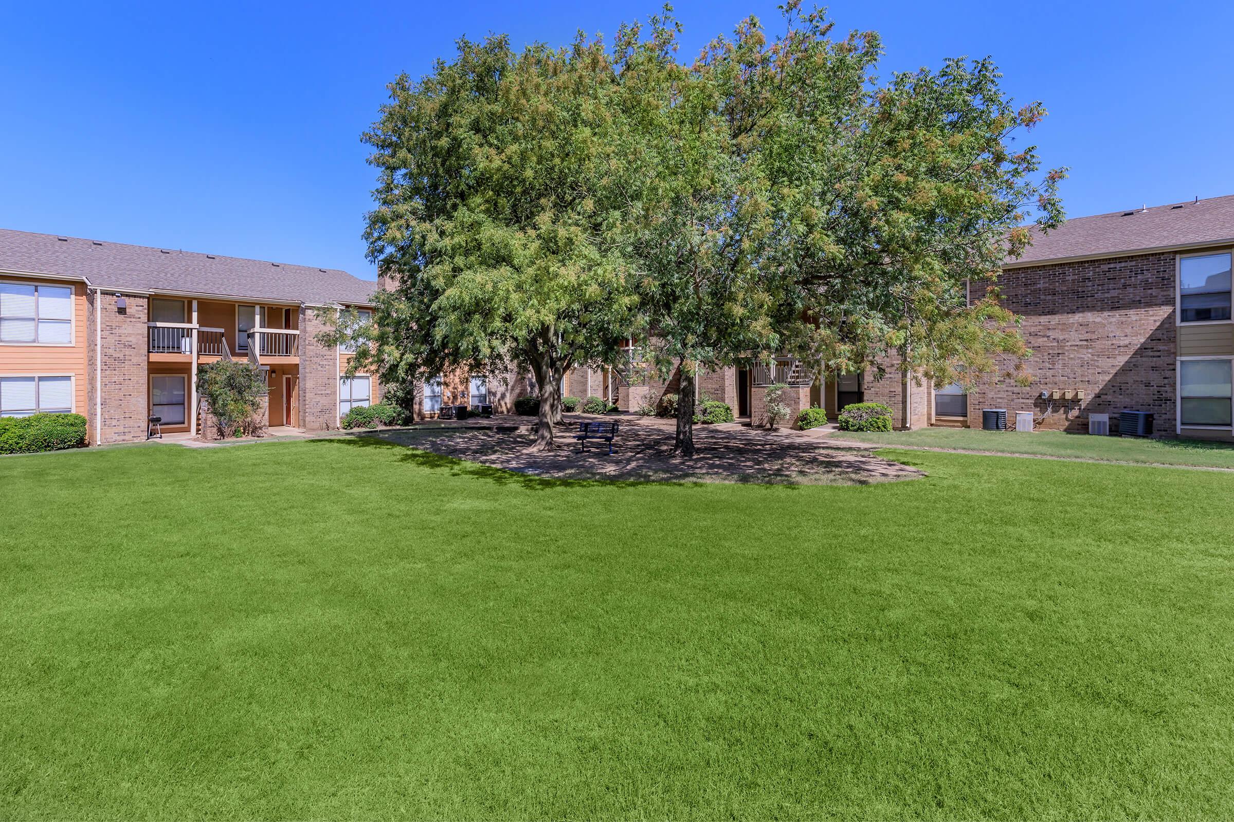 A green lawn surrounded by two-story brick apartment buildings. A large tree provides shade in the center of the grassy area, with benches underneath. The clear blue sky adds to the peaceful atmosphere of the outdoor space.